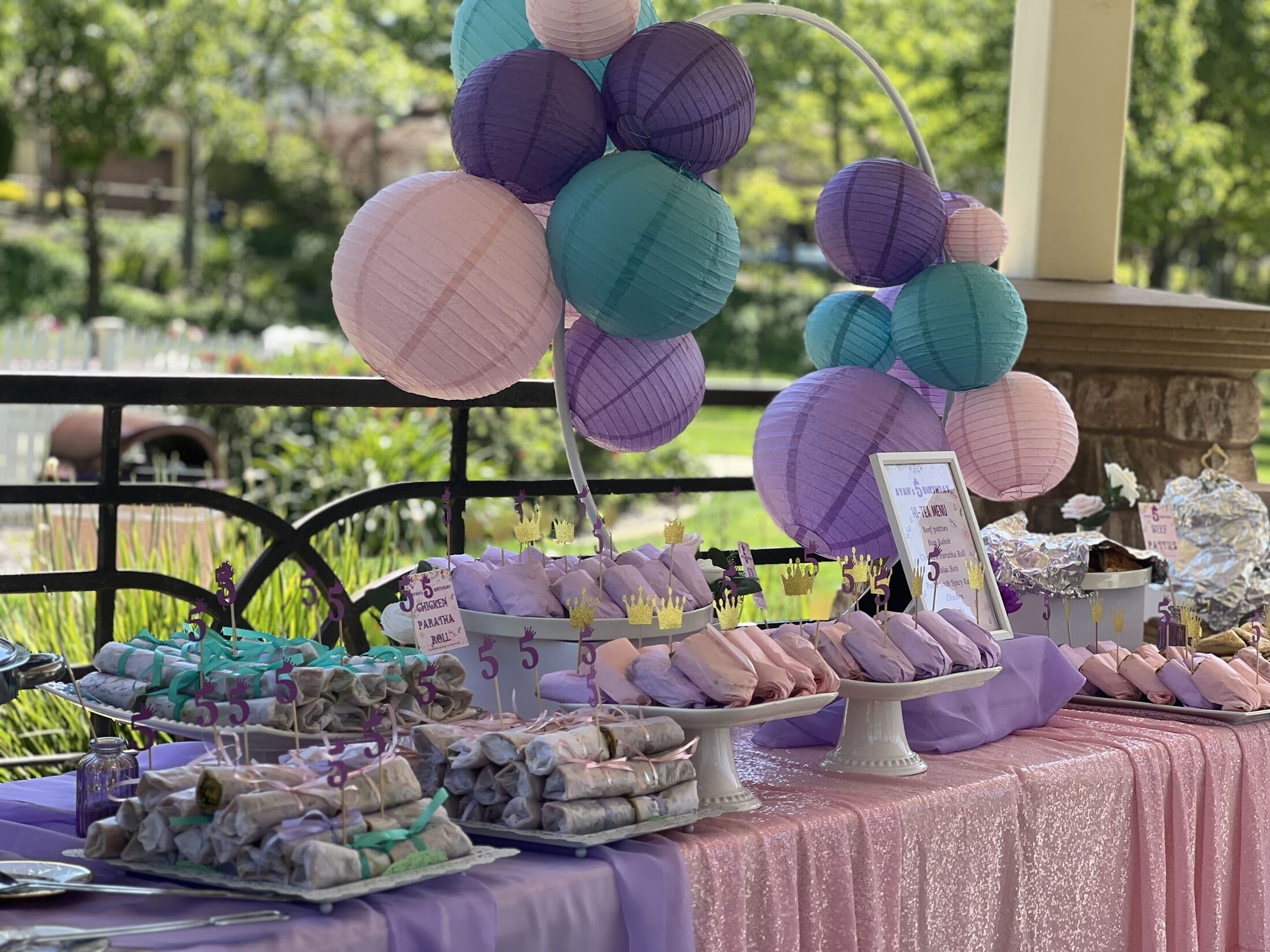 An outdoor party food table dressed in pink and lavender, with paper-wrapped wrap rolls on white stands beneath a cluster of purple and teal paper lanterns.
