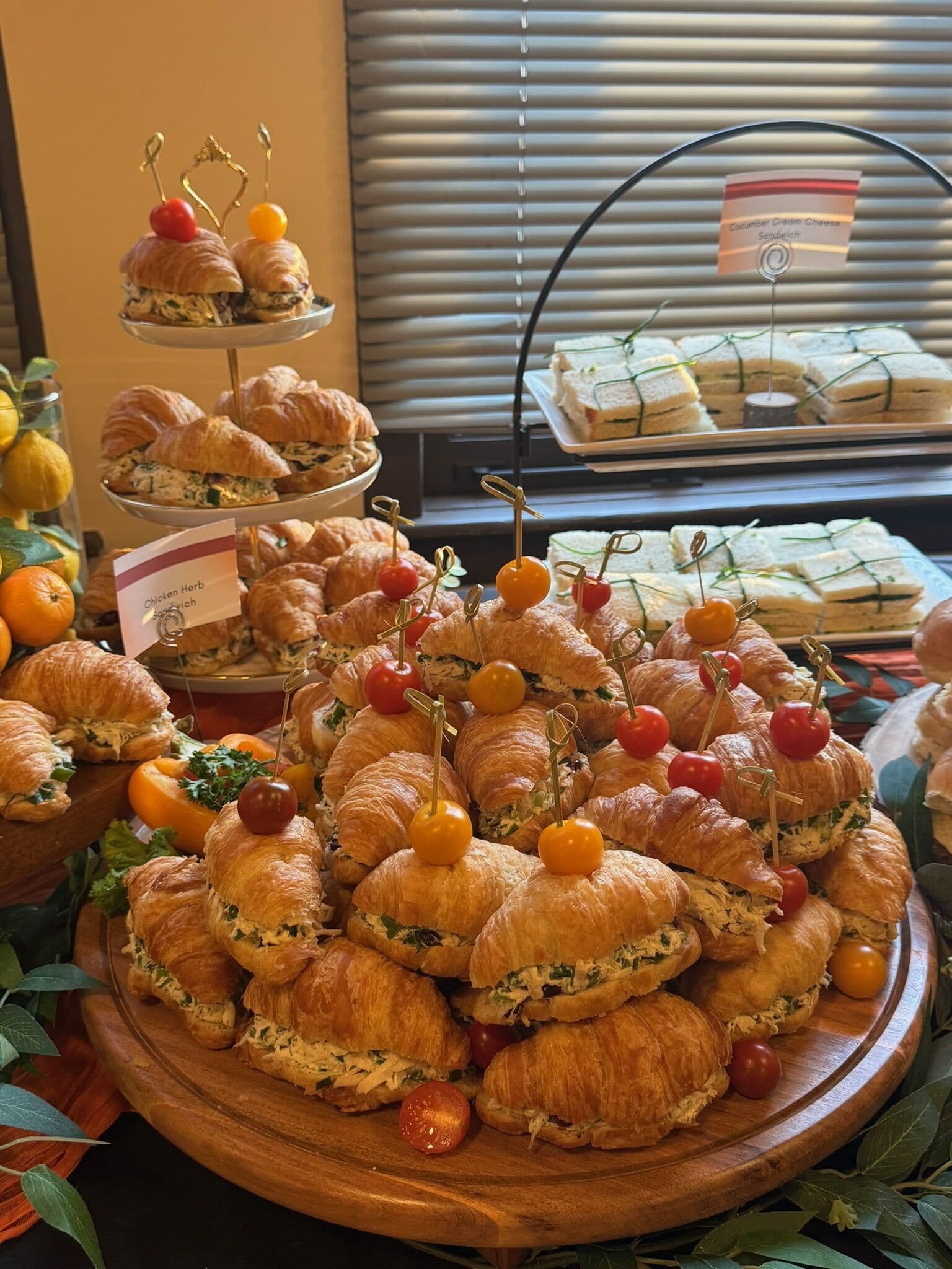 A wooden board mounded with croissant sandwiches filled with herbed chicken salad and pinned with red and yellow cherry tomatoes, with more croissants and tea sandwiches tiered behind for a Hi-Tea.