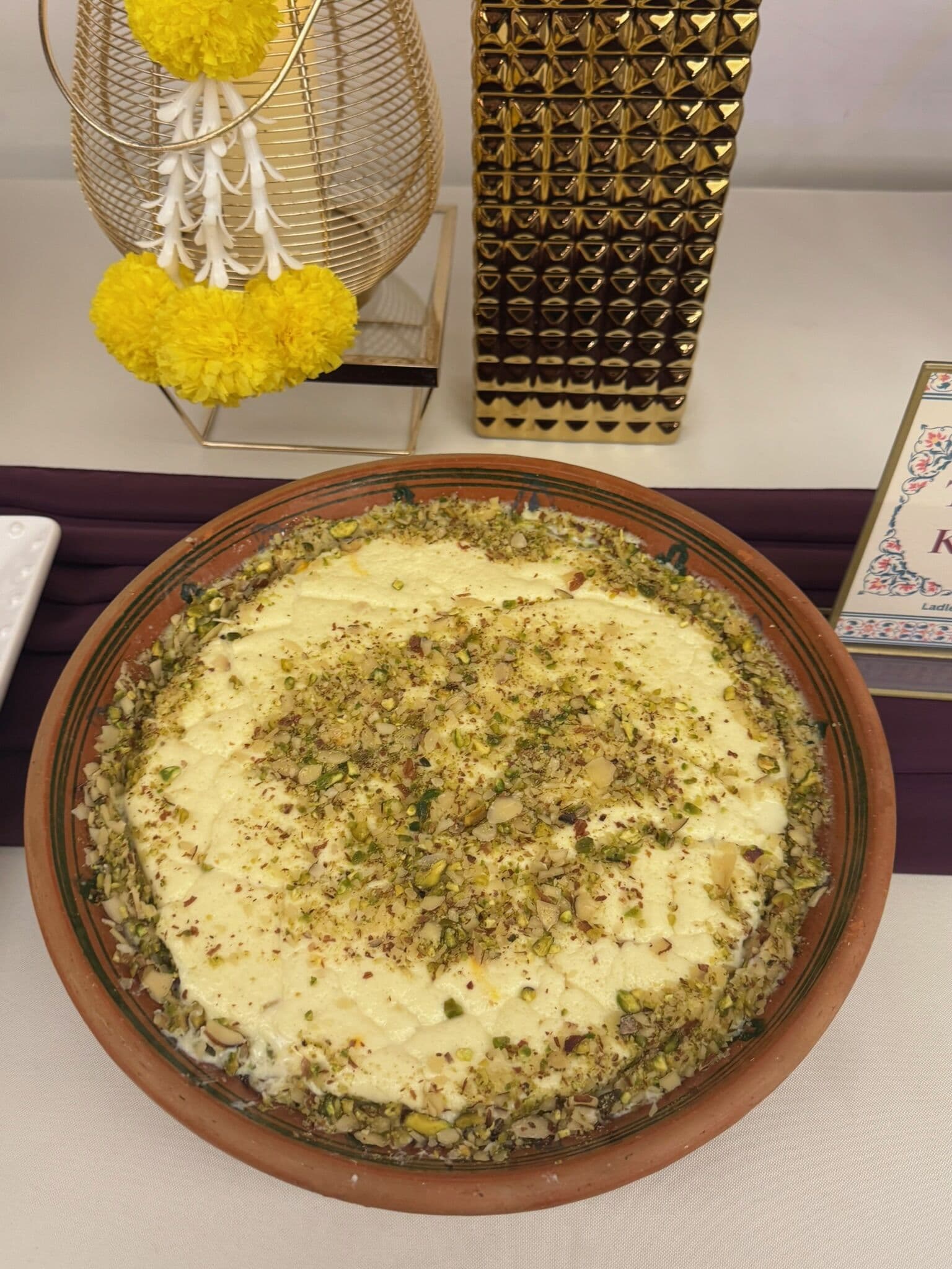 A terracotta dish of set milk dessert, likely a halal cream-topped sheer or custard, edged and dusted with chopped pistachio, styled with marigold and gold accents.