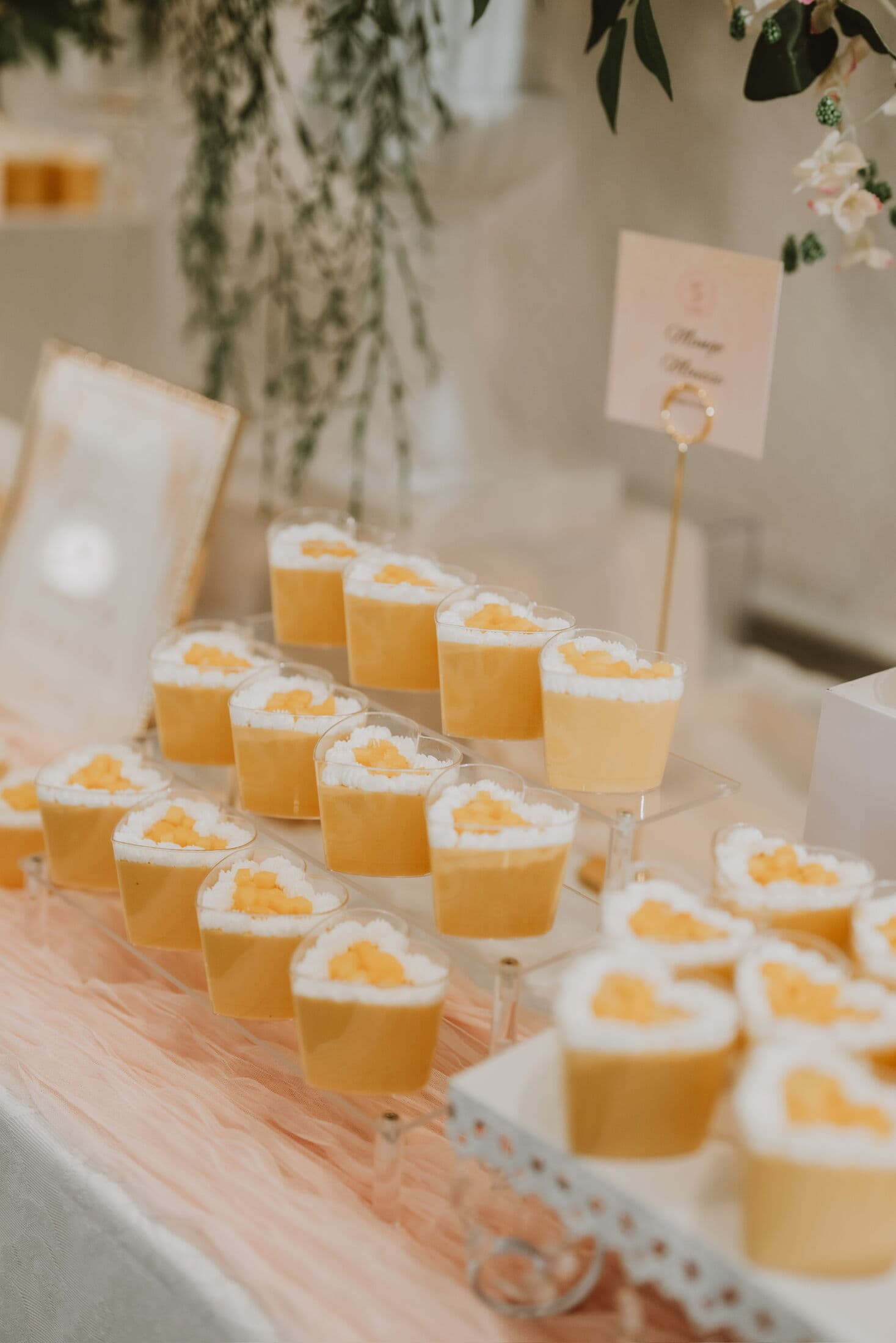 Rows of individual mango mousse cups topped with piped cream and diced mango, arranged on tiered acrylic risers over a blush runner with trailing greenery at a Bay Area event.
