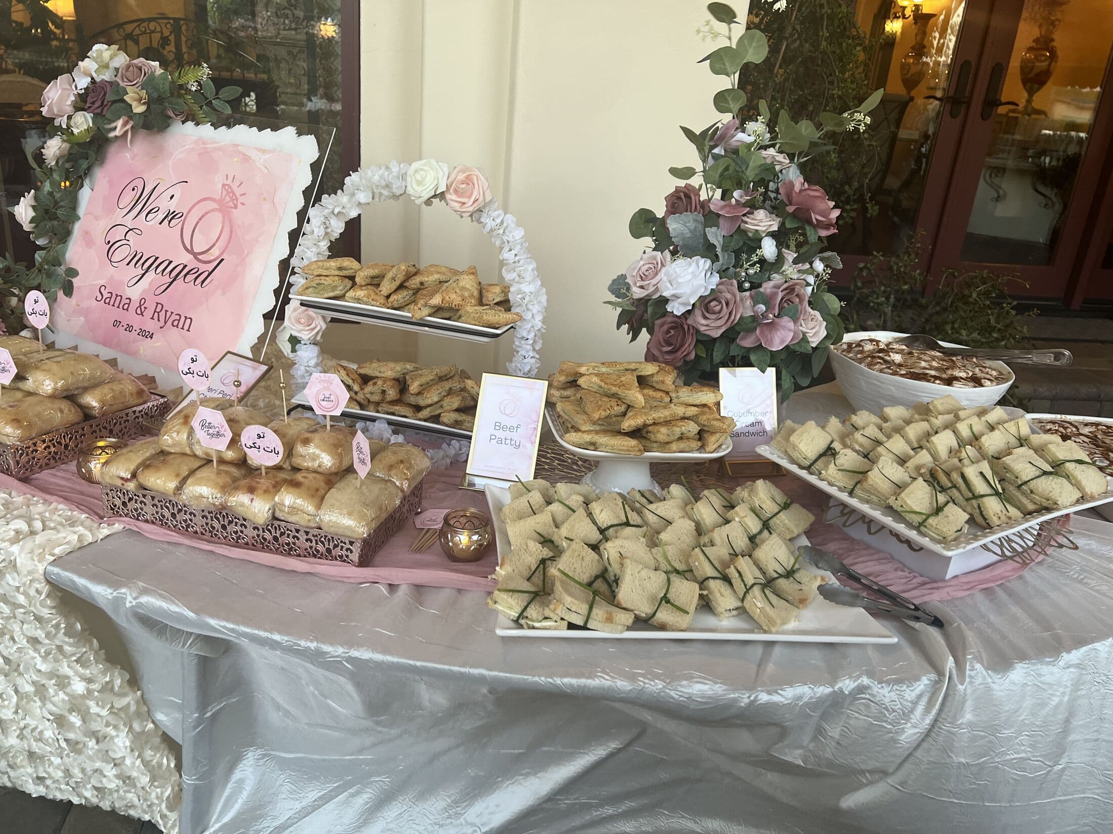 Outdoor engagement grazing table with a pink We're Engaged sign, layered platters of samosas, beef patties, finger sandwiches and pastries with labeled cards and blush rose arrangements.
