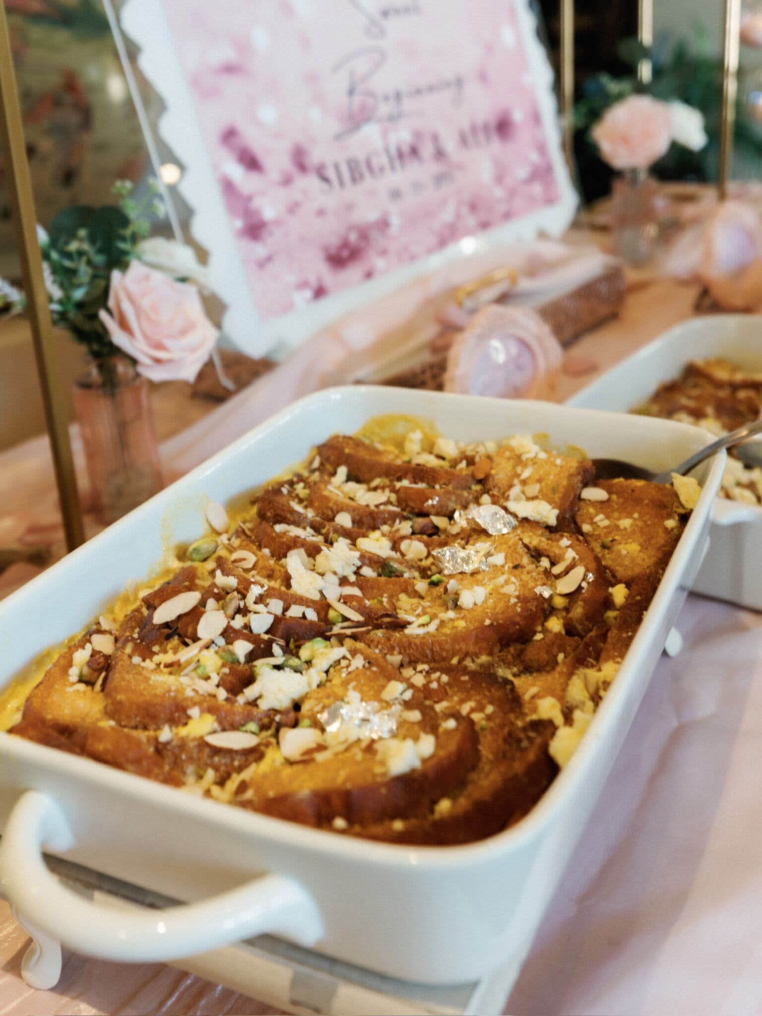 White handled baking dish of halal shahi tukra bread pudding in saffron custard, topped with slivered almonds, pistachios and silver leaf, set beside a pink Sweet Beginnings sign and roses.