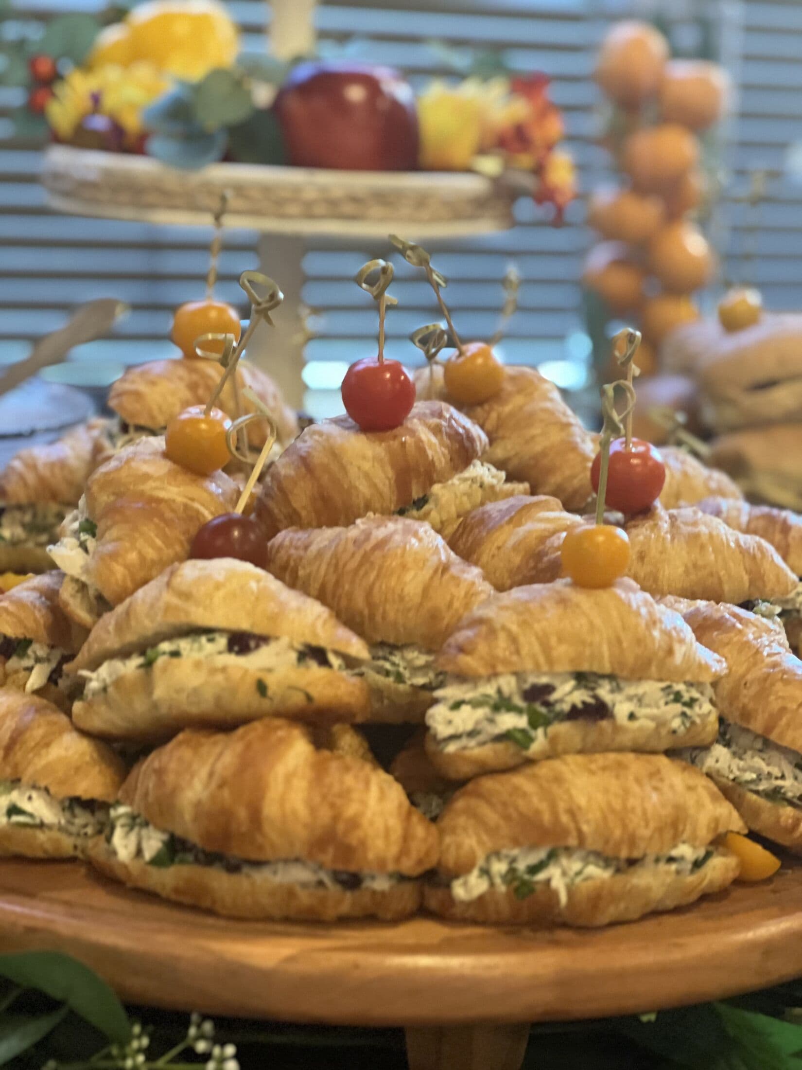 Wooden cake stand piled with croissant tea sandwiches filled with herbed chicken salad and cranberries, topped with cherry tomatoes on decorative picks, from the Ladle n Wok Hi-Tea spread.