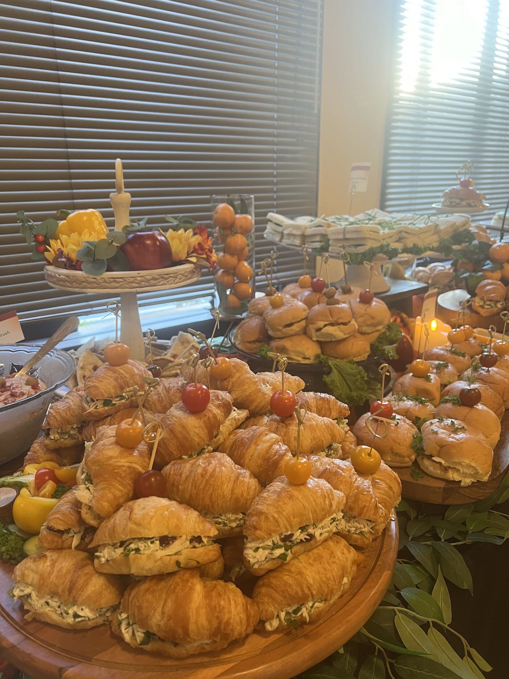 Indoor Hi-Tea table layered with wooden platters of croissant chicken salad sandwiches and finger sandwiches, accented with cherry tomato picks, candlelight and a fruit-topped cake stand.
