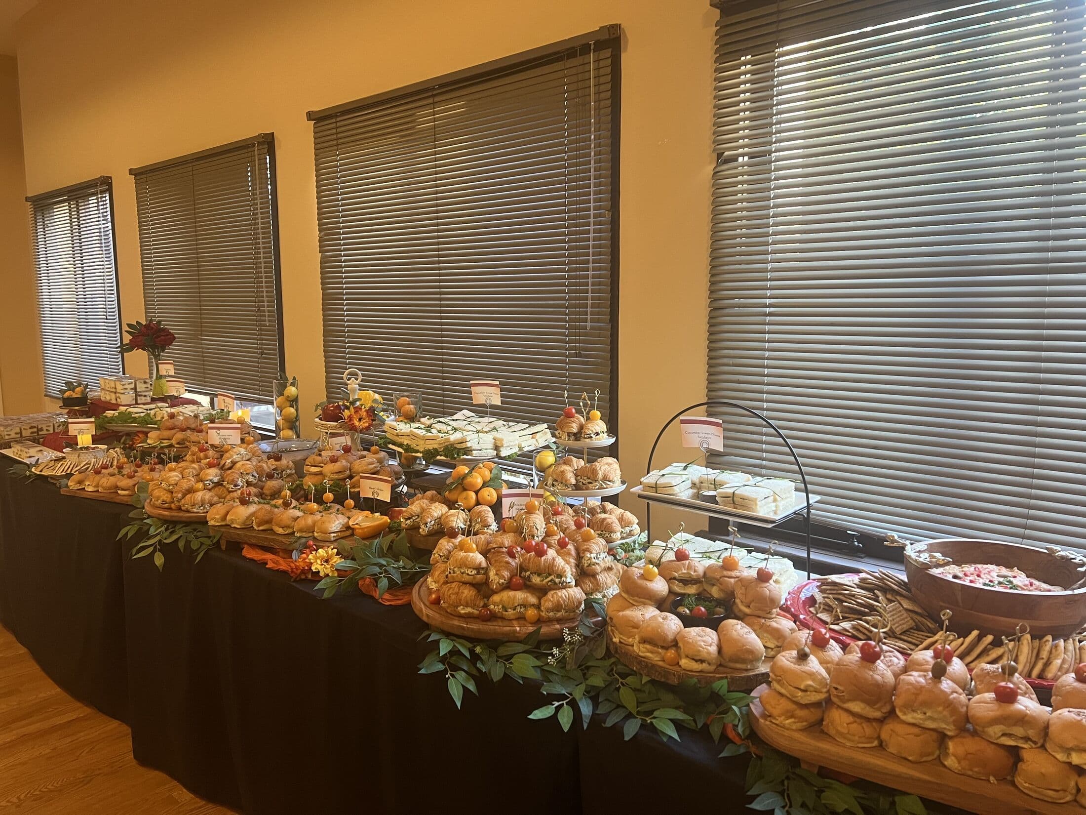 Long banquet grazing spread against blind-covered windows, with tiered platters of pastries, sliders, sandwiches and desserts trailing greenery down a black-linen table in a Bay Area venue.