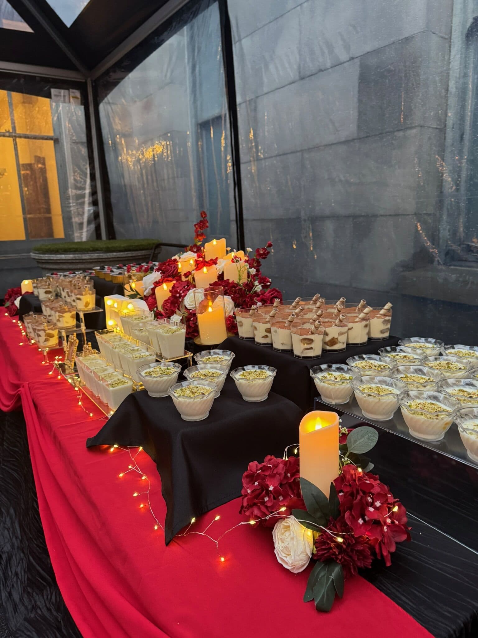 Nighttime dessert station with long rows of individual trifle and pistachio-topped pudding cups on black linen, lit by pillar candles, fairy lights and deep-red floral accents.