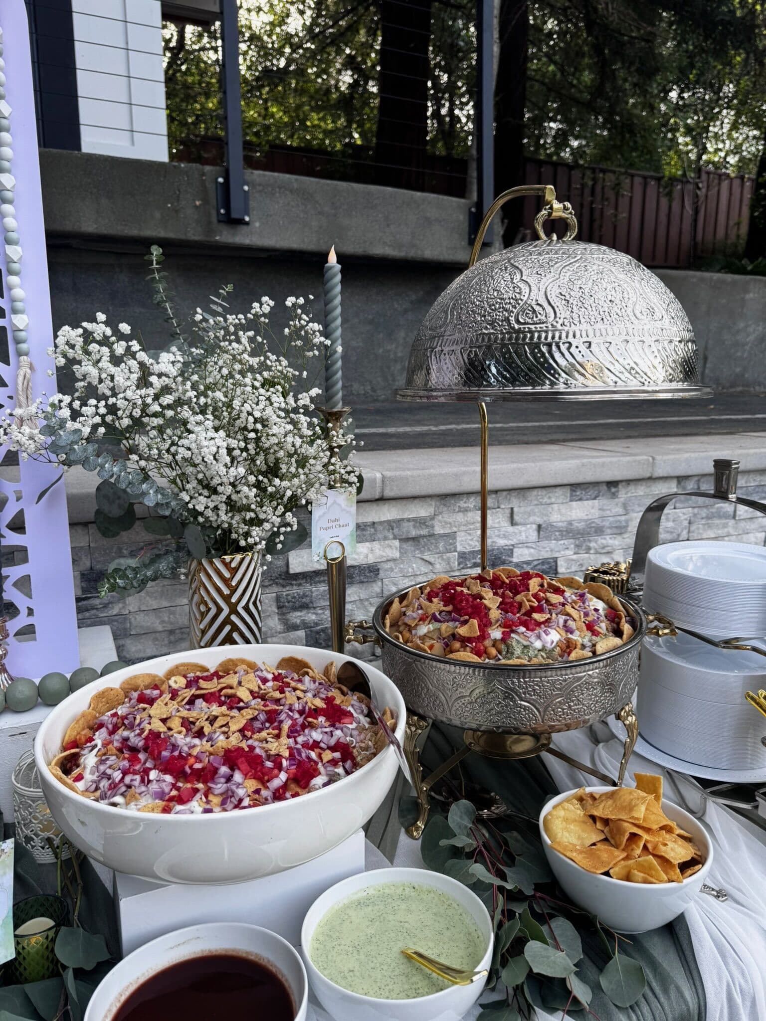 Outdoor chaat station with a large white bowl and an ornate silver chafing dish of dahi bhalla or fruit chaat garnished with pomegranate and nuts, served with tortilla chips, mint chutney and tamarind sauce.