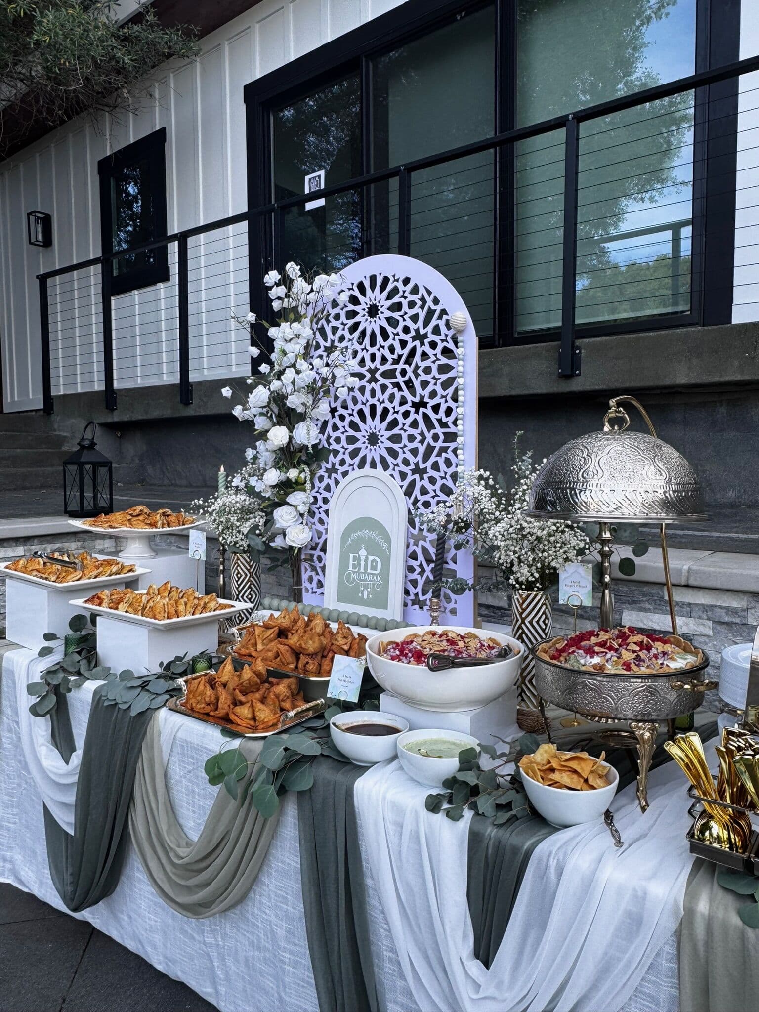Styled outdoor buffet before a white Eid-themed arch and baby's breath florals, with white bowls of samosas, chaat and chips alongside a domed silver chafing dish and gold serving tongs.