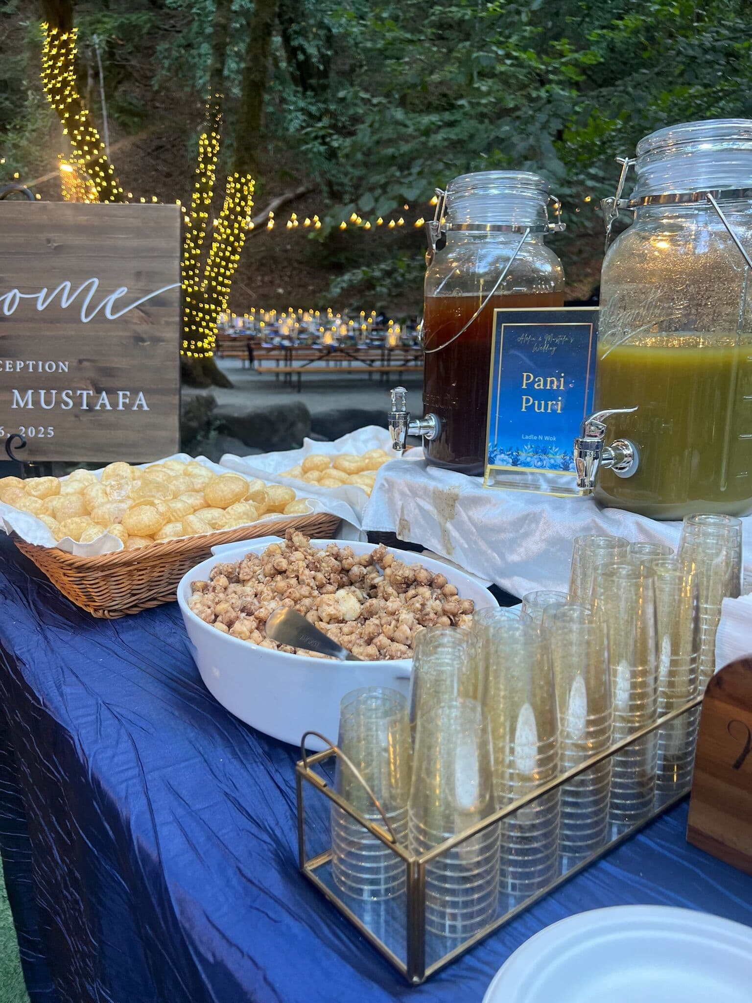 Evening pani puri station under string lights, with a basket of crisp puri shells, a bowl of masala filling, tall gold-rimmed cups and glass dispensers of tamarind and mint waters marked by a Pani Puri card.