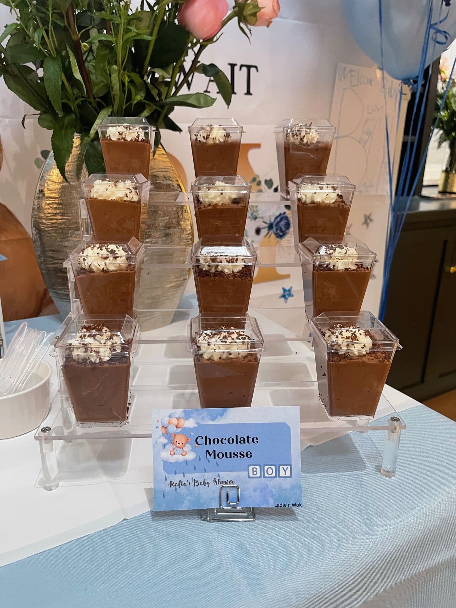Tiered acrylic display of Chocolate Mousse cups topped with cream and shaved chocolate at a blue-themed baby shower, with a Chocolate Mousse card and roses behind.
