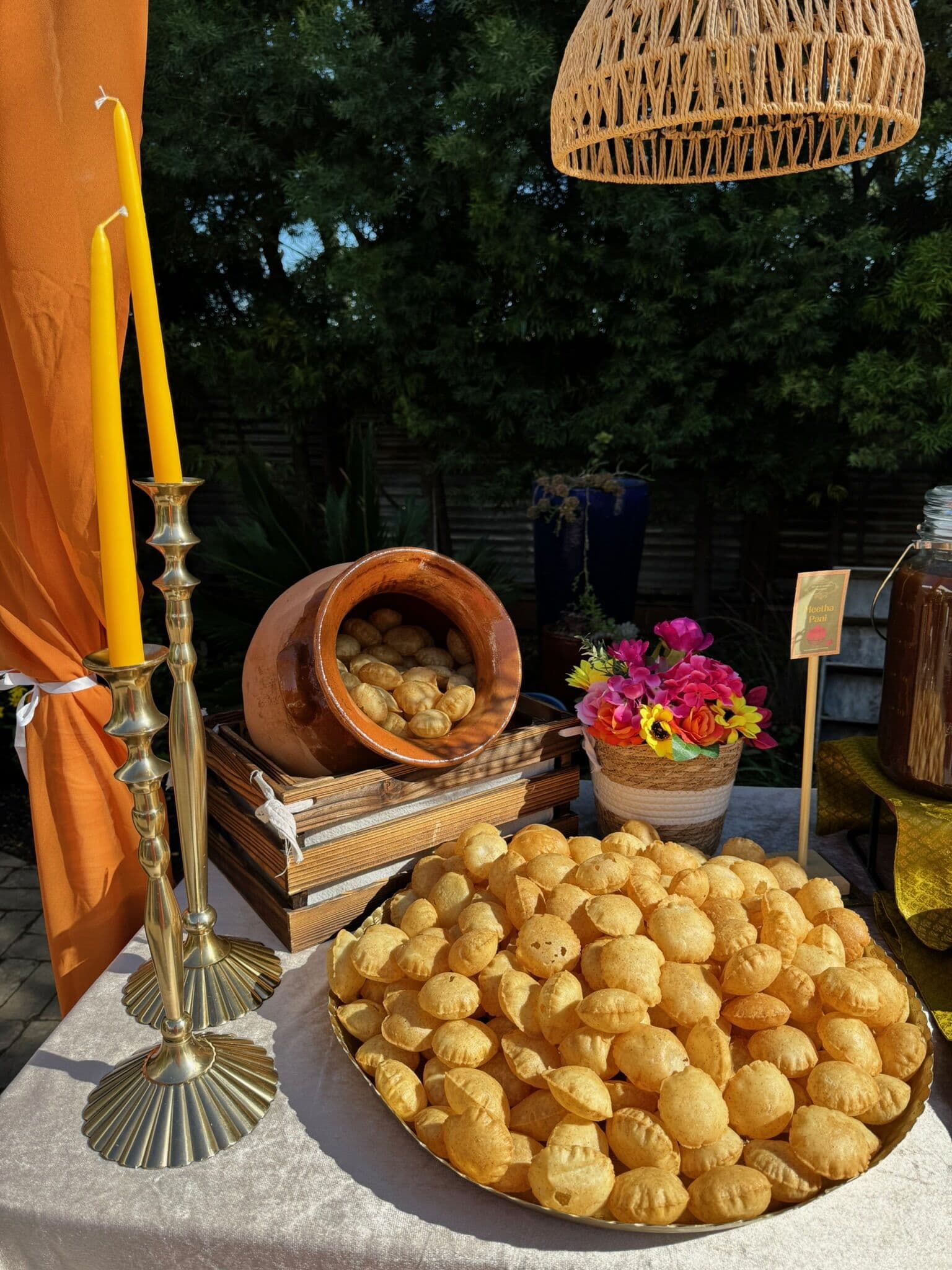 Golden tray heaped with crisp puri shells for pani puri on a rustic outdoor table, styled with tall brass candlesticks, stacked books, a bright floral pot and a beverage dispenser at dusk.