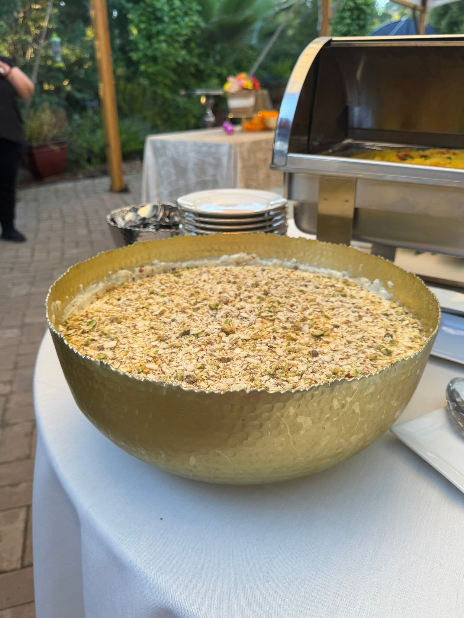 Large hammered gold bowl of kheer or seviyan dessert dusted with crushed pistachios and almonds on a linen-draped table, with stacked plates and a silver roll-top chafer of rice behind.