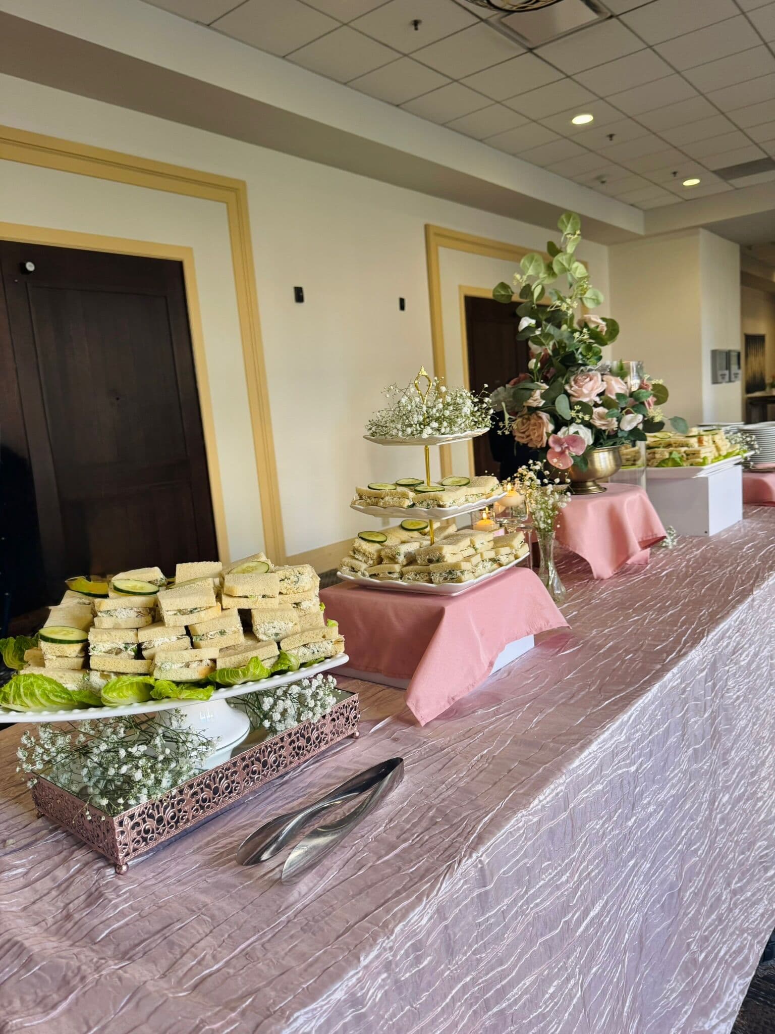 Indoor Hi-Tea table with tiered stands and platters of cucumber and herbed finger sandwiches on lettuce, dressed with blush linens, baby's breath and a rose-and-eucalyptus centerpiece.