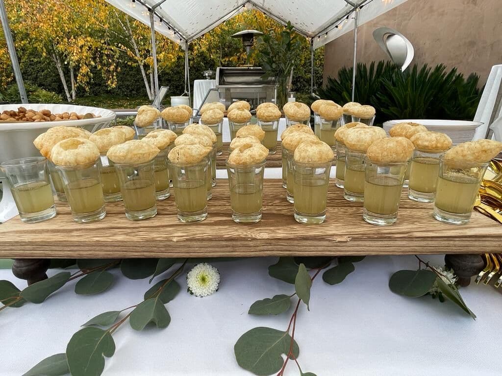 Golgappa shot station on a rustic wood plank under a marquee, with tall glasses of spiced pani each capped by a crisp puri shell, styled with trailing eucalyptus and a white bloom.