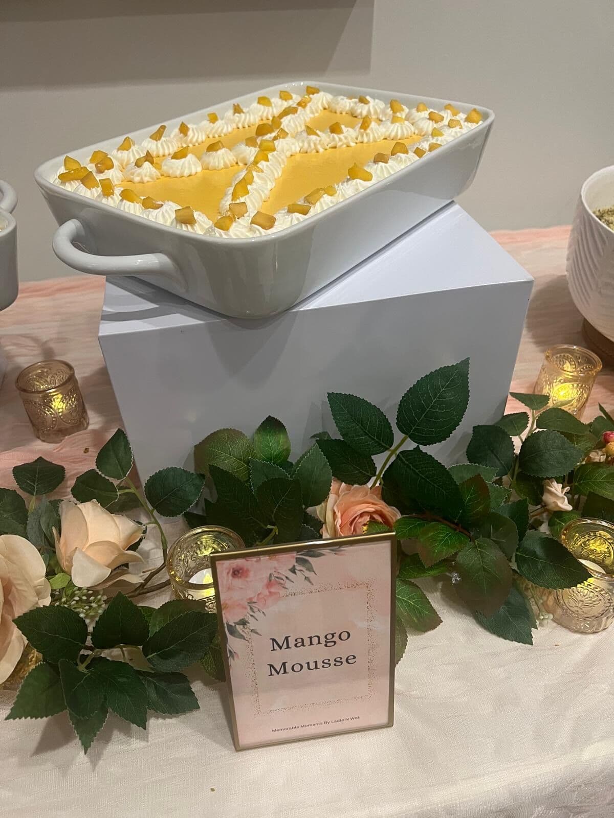 White handled dish of Mango Mousse piped with cream rosettes and diced mango on a raised riser, framed by ivory roses, greenery and votives beside a floral Mango Mousse card.