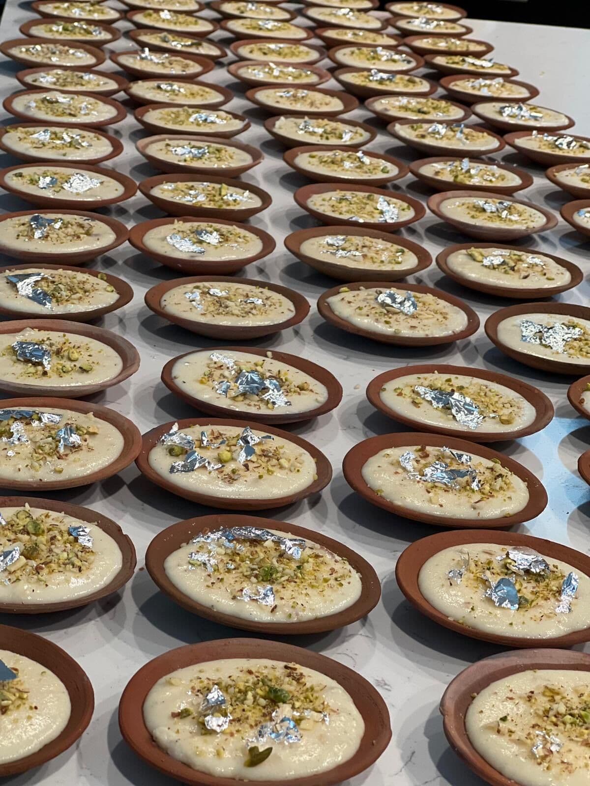 Dozens of terracotta clay dishes of kheer rice pudding lined in neat rows, each garnished with crushed pistachios and delicate silver leaf, prepped in the Ladle n Wok kitchen.