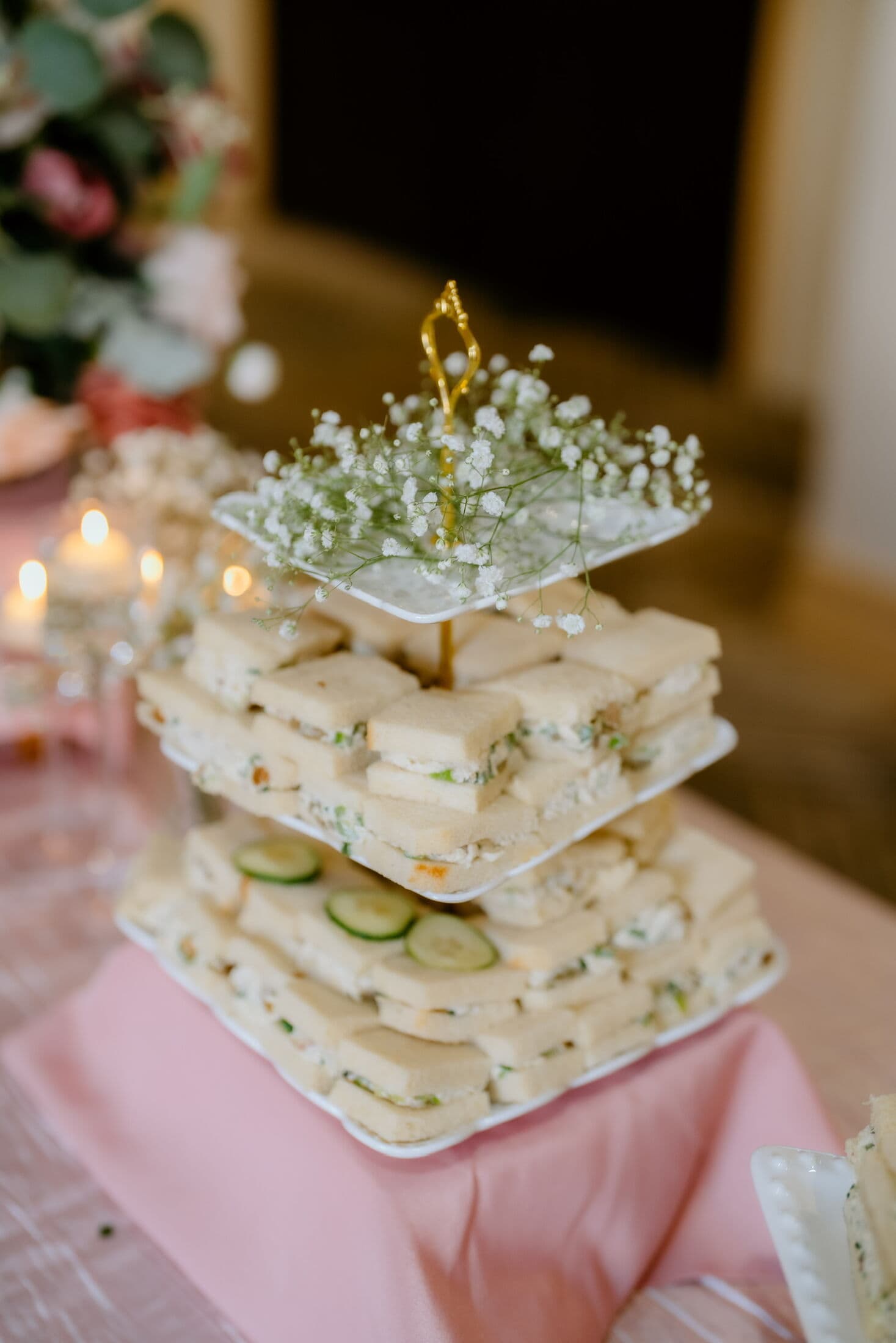 Three-tier square stand of crustless finger sandwiches filled with herbed chicken and cucumber, topped with cucumber slices and baby's breath, on a blush linen Hi-Tea table with candlelight.