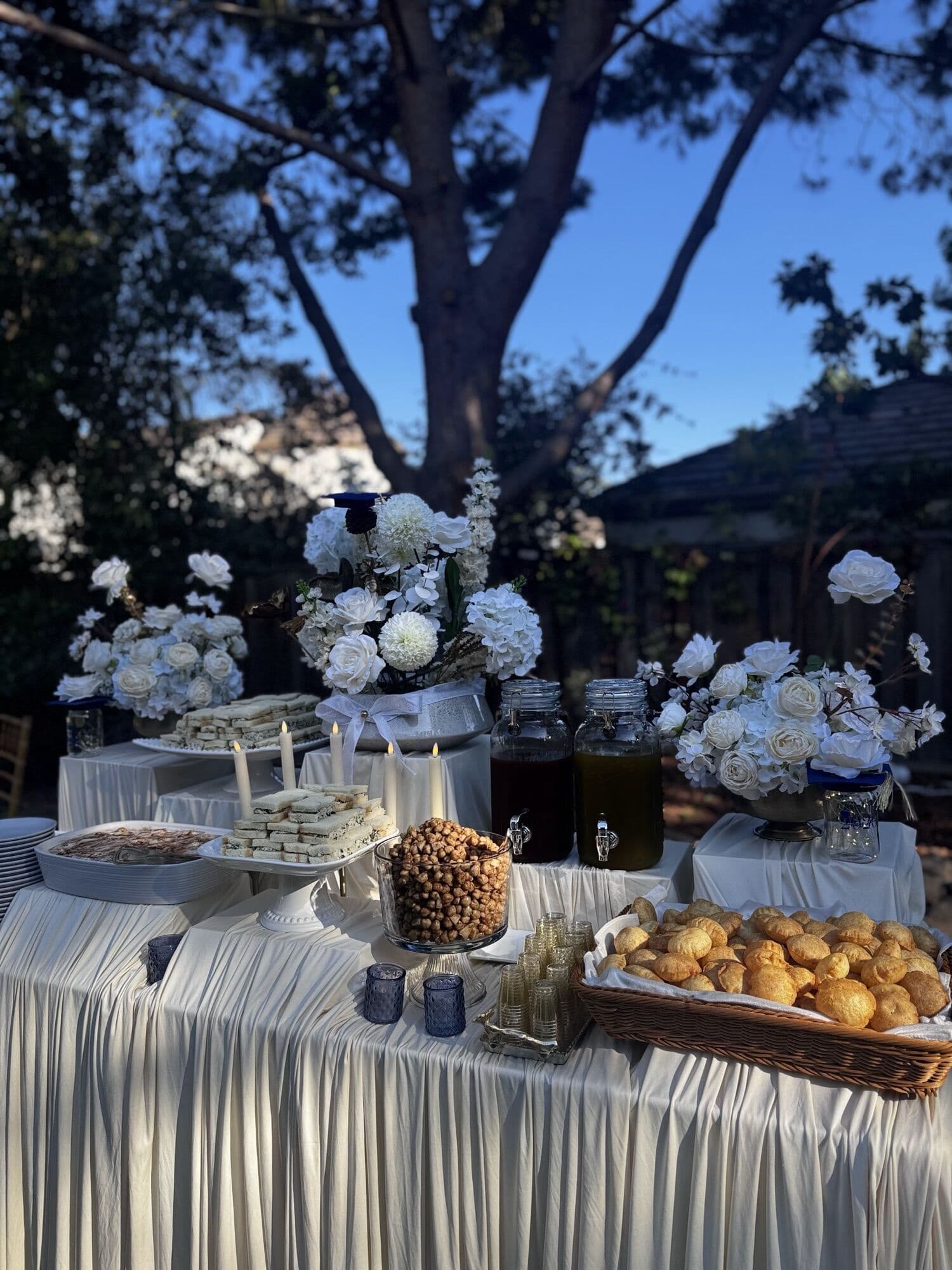 Garden buffet under a tree at golden hour — tiers of finger sandwiches, a glass bowl of spiced chickpeas, glass dispensers of tamarind and mint waters, a basket of crisp puri shells and tall white floral arrangements on ivory draped tables.