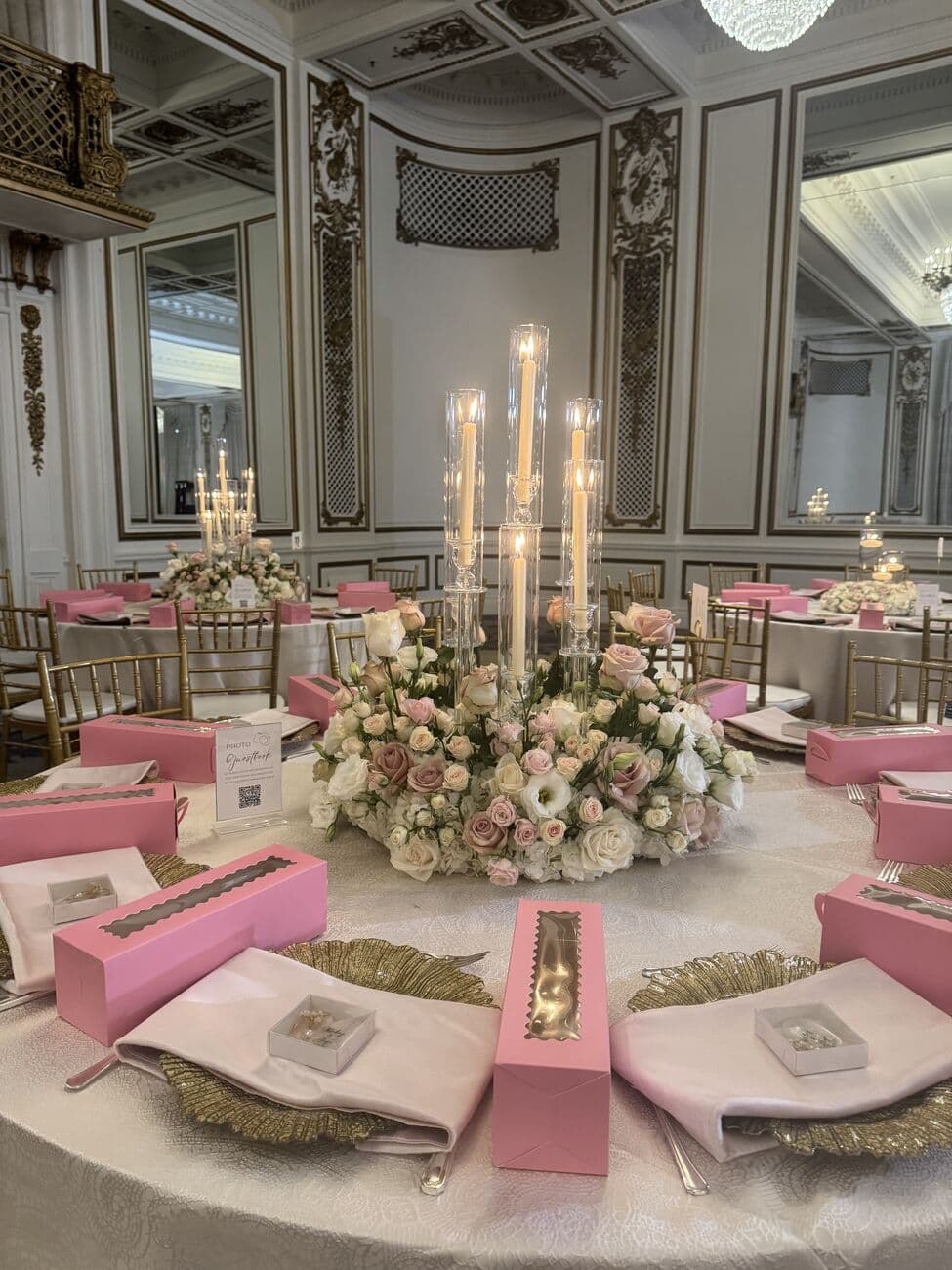 Ornate ballroom round table with a low blush rose centerpiece ringed by crystal candelabras, gold filigree chargers, blush napkins and pink favour boxes, set beneath gilded panelling and mirrors.