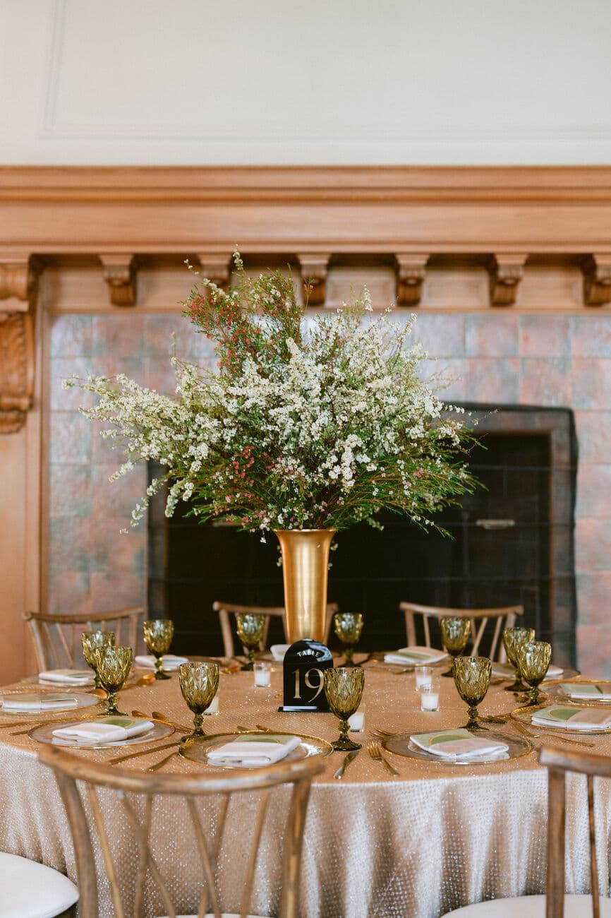 Round guest table before a carved wood and tiled fireplace, set with a tall brass urn of white blossom and berried greenery, gold-rimmed chargers, smoked-green goblets and a black table number.