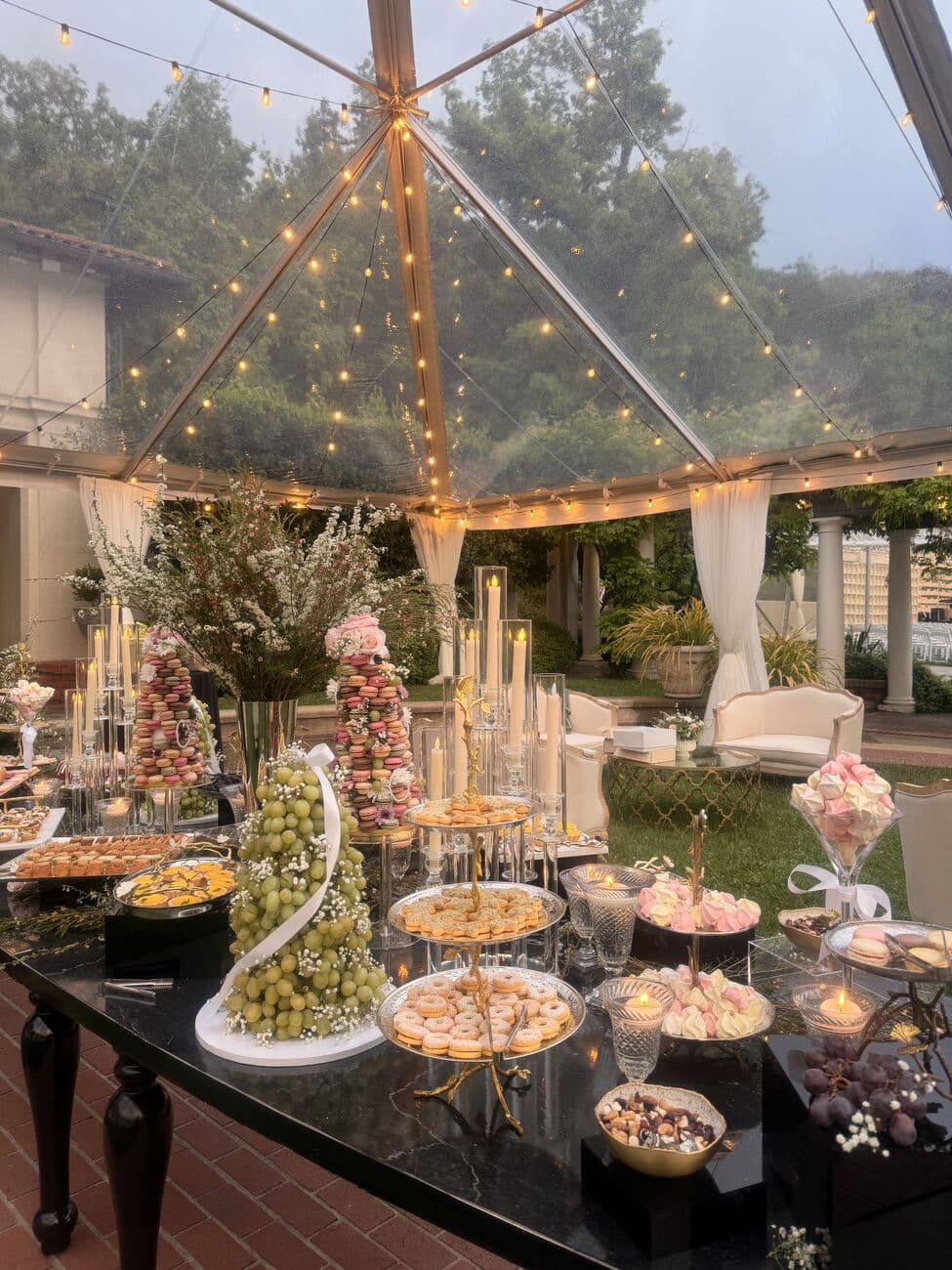 Dessert table under a clear tented marquee strung with fairy lights, with macaron towers, a cascading green grape display and tiered trays of pastries and meringues on black marble at dusk.