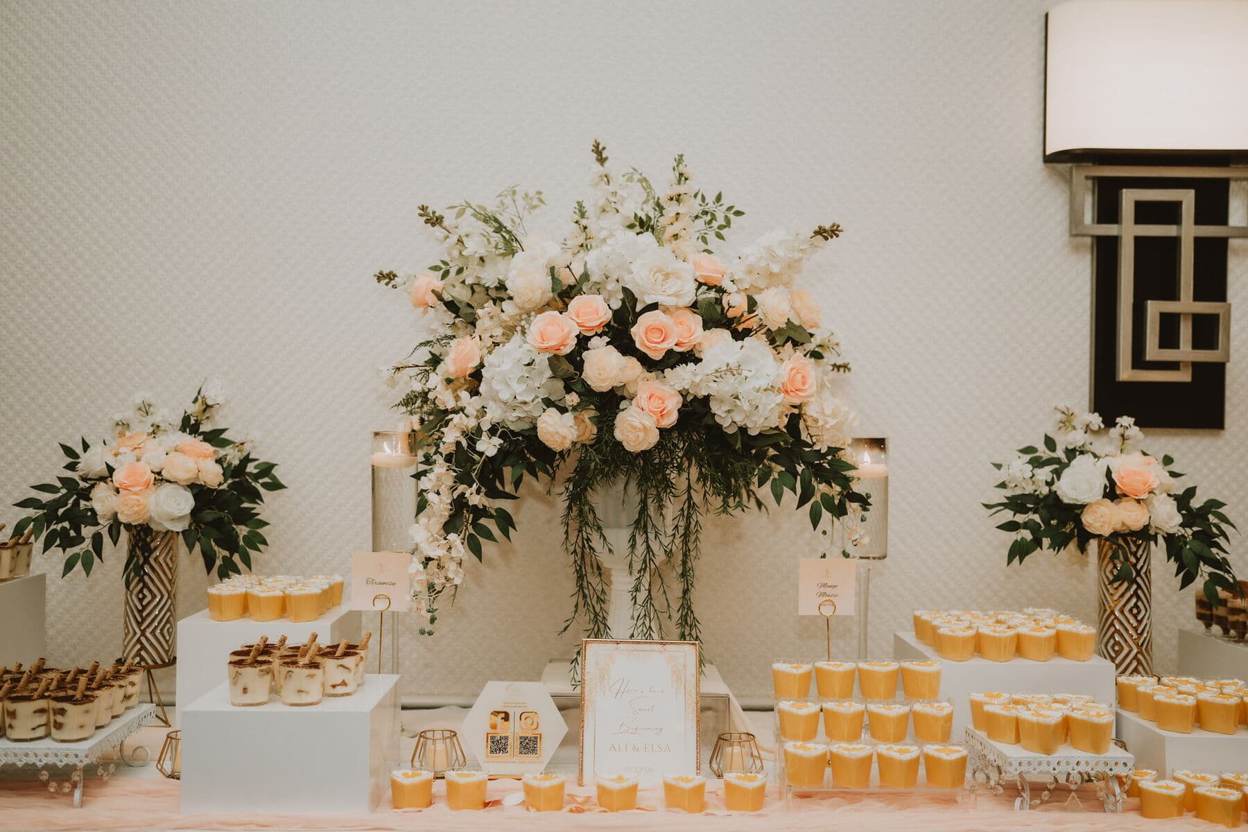 Dessert display against a textured ivory wall with a tall peach-and-white floral centerpiece, tiered white risers of mousse cups in gold sleeves, tiramisu portions and a printed welcome card.