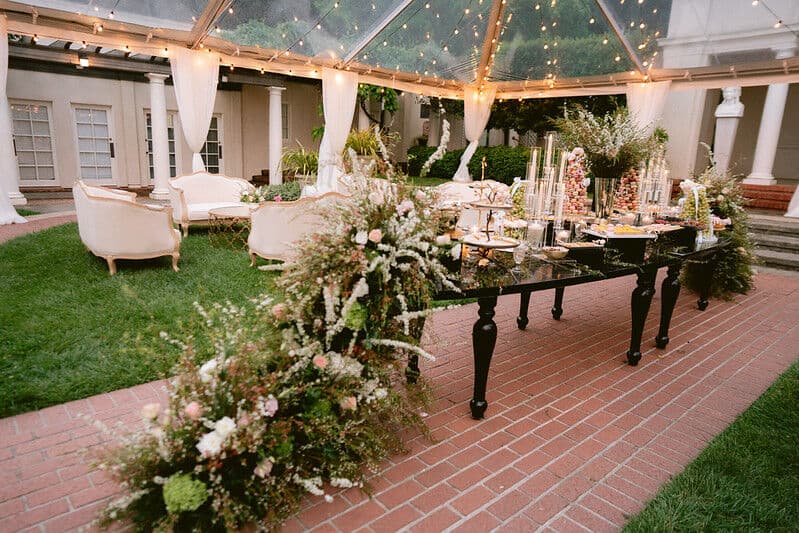 Clear-tented garden lounge at dusk with cream settees on the lawn, a black marble dessert table of tiered sweets and tall tapers, and a long bed of blush and ivory florals along the brick path.