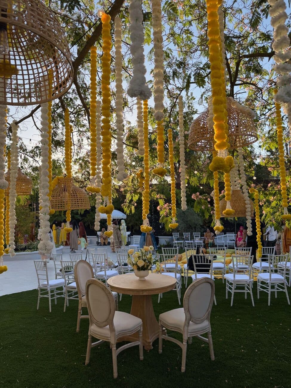 Outdoor haldi under a tree hung with strands of yellow and white marigold garlands and woven rattan pendant lanterns, over white Chiavari chairs and a round table with a yellow rose centerpiece.