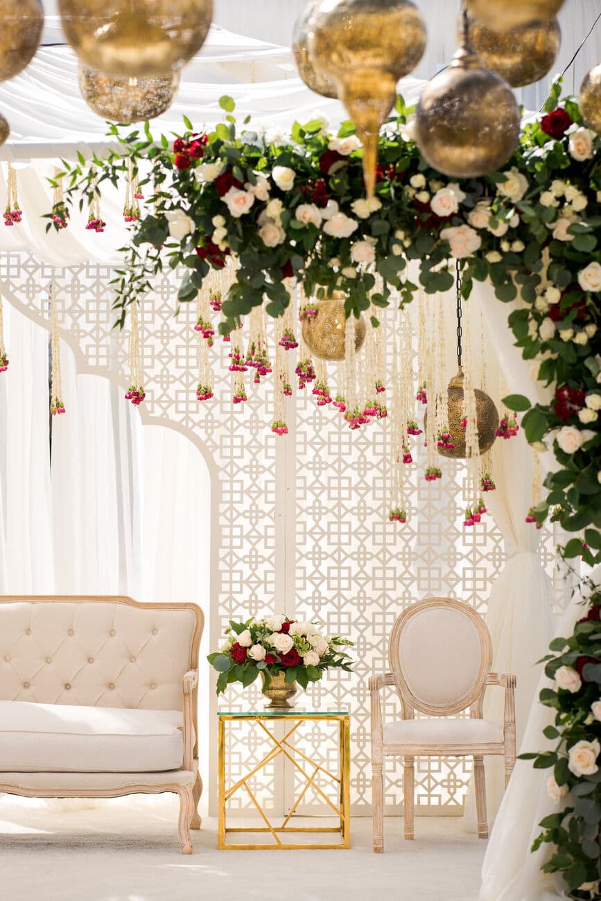Detail of a white mandap — a canopy of burgundy and ivory roses with hanging marigold strands and gold lanterns over a lattice screen, above a tufted cream settee and a gold side table.