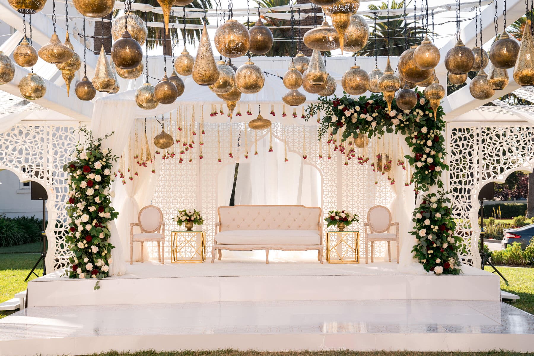 Outdoor white mandap under a canopy of hanging gold mercury-glass lanterns, with lattice screens, garlands of burgundy and ivory roses, a tufted cream settee and gold side tables on a white platform.