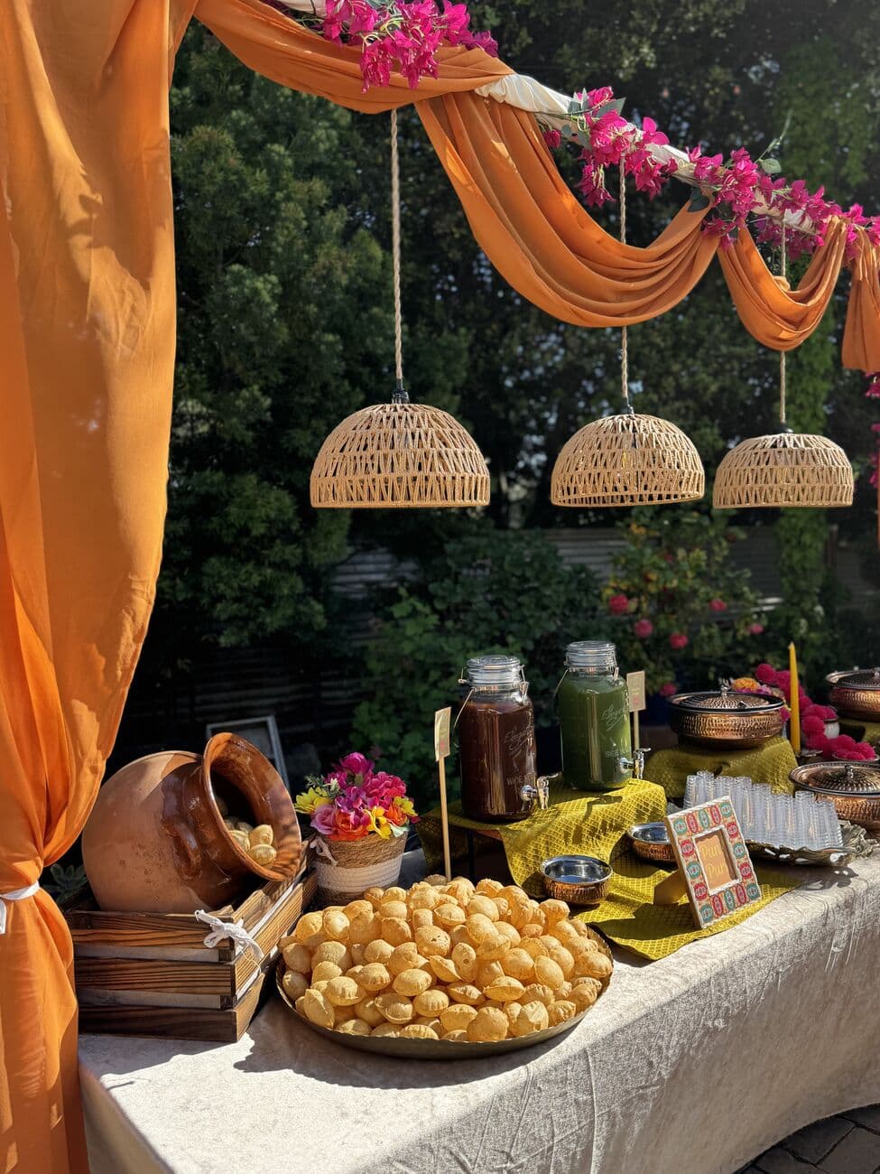 Pani puri station under orange chiffon drapery and bougainvillea, with woven rattan pendant lamps, a brass platter of puri shells, glass dispensers of tamarind and mint water and copper serving pots.