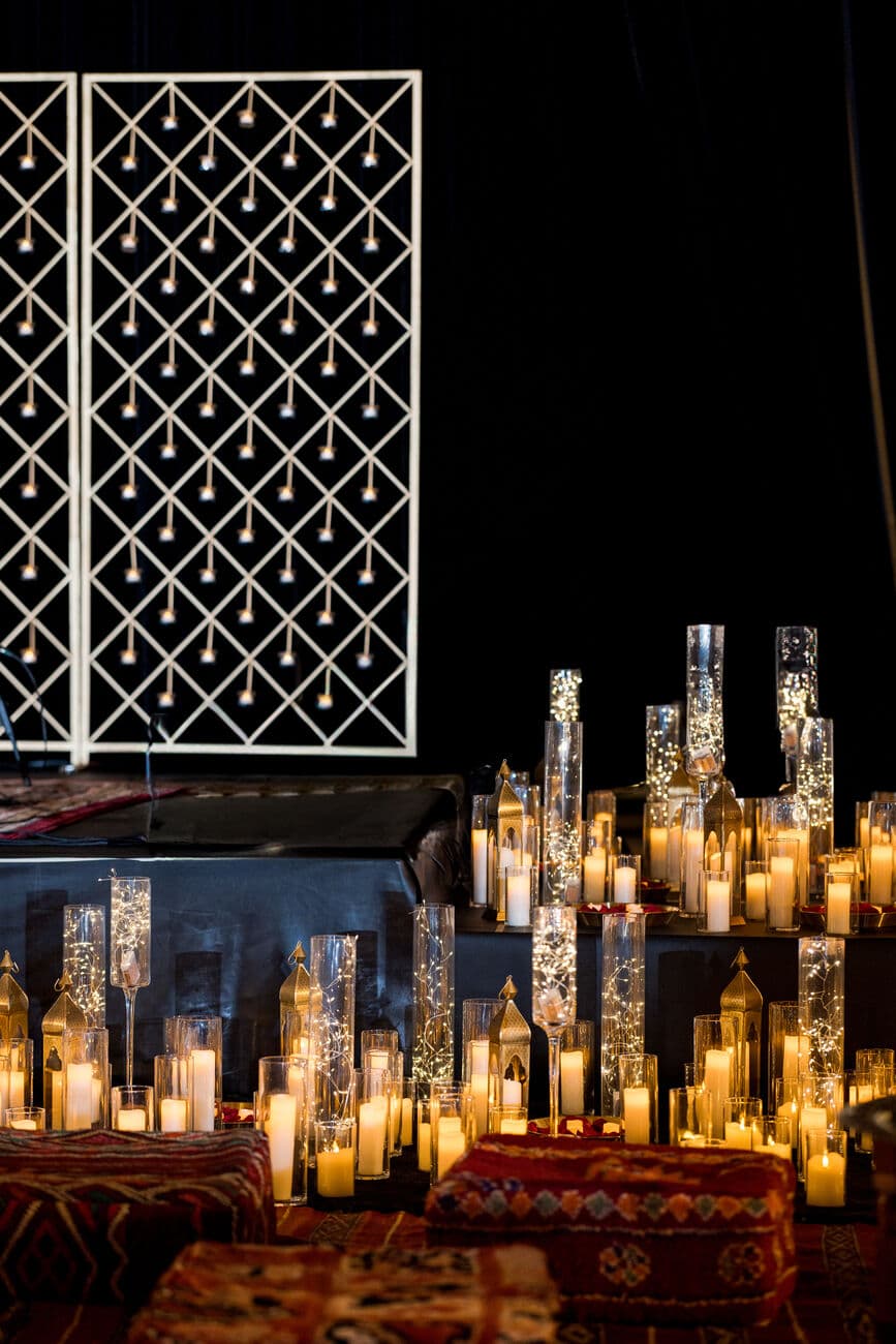 Nighttime qawali setting with two white lattice screens strung with fairy lights above a low stage, surrounded by dozens of pillar candles in glass cylinders, brass lanterns and patterned floor cushions.