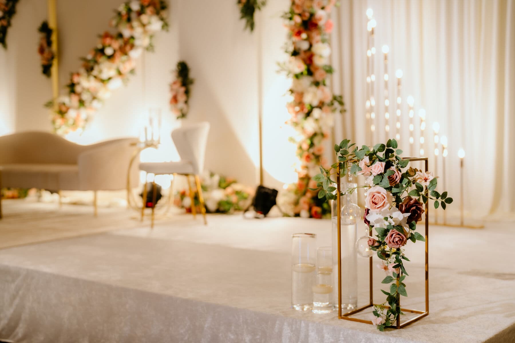 Ivory wedding stage with cascading garlands of peach, burgundy and white roses down white columns, a cream settee and accent chair, and a gold frame centerpiece of roses and eucalyptus.