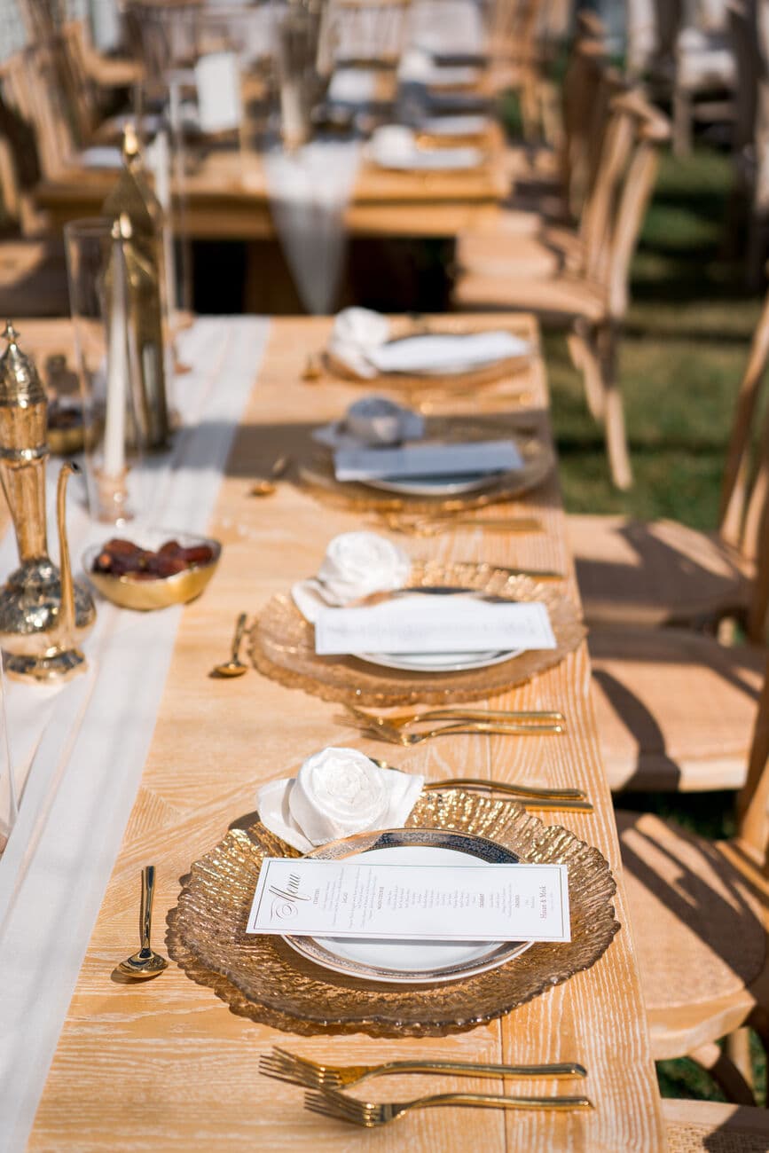 Long outdoor wooden banquet table set with gold textured chargers, white rose napkin folds, printed menus, brass flatware and an engraved brass dallah beside a bowl of dates.