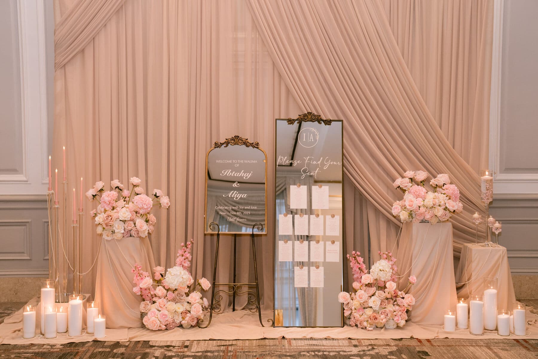 Blush pleated drapery backdrop with a gold-framed welcome mirror and a 'Please Find Your Seat' seating chart on easels, flanked by pedestals of blush and ivory roses and banks of white pillar candles.