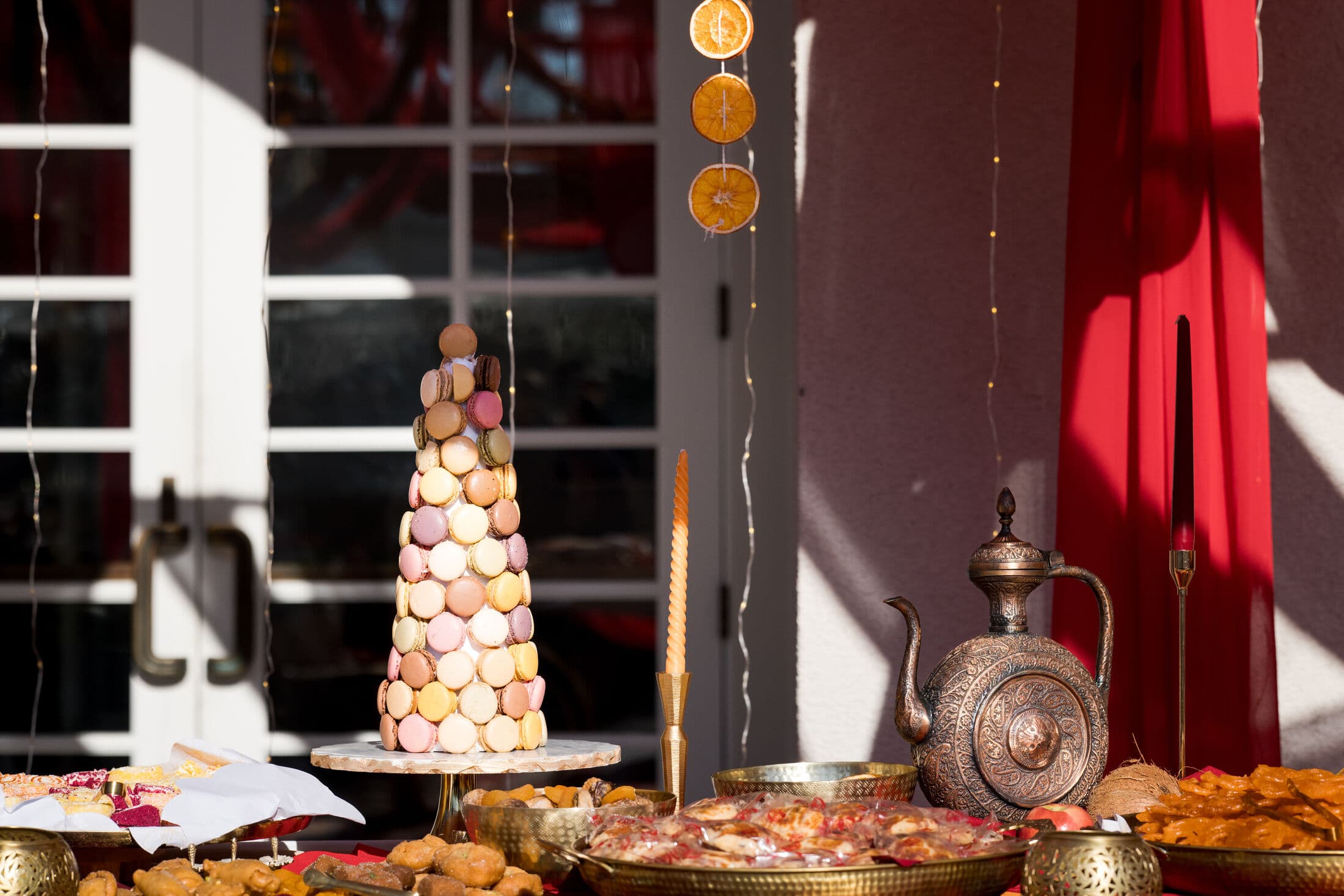 A pastel macaron tower on a marble cake stand anchors a sunlit Bay Area dessert table, surrounded by trays of golden fried sweets, an engraved copper teapot, and hanging dried orange slices against red draping.