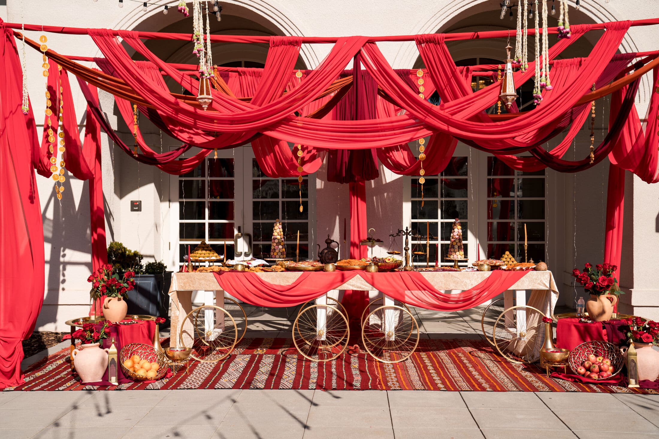 An outdoor dessert setup by Memorable Moments by Ladle n Wok with white gold-wheeled cart tables under crimson ceiling draping, dressed with red runners, floral urns, and platters of sweets over a patterned rug.