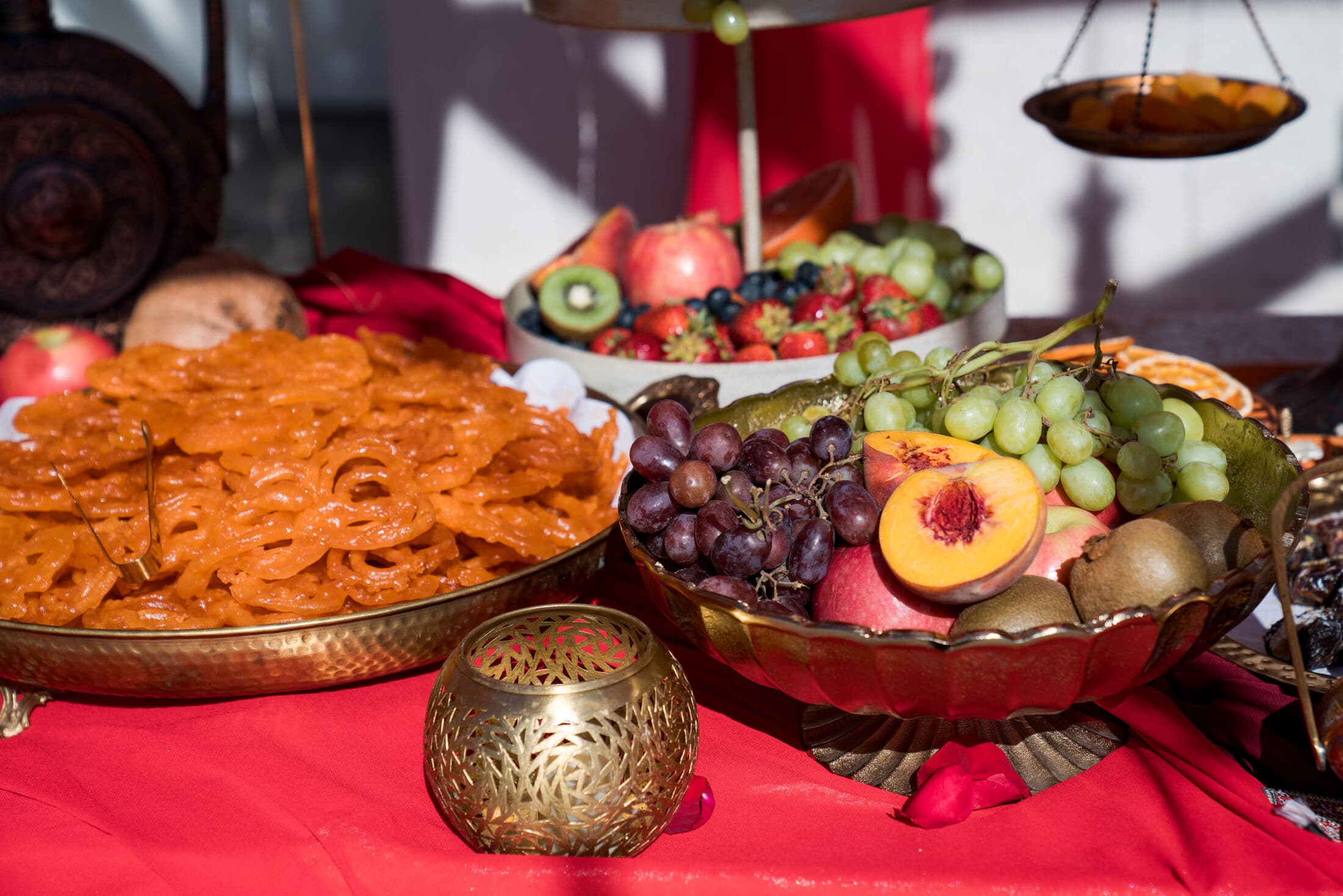 A brass tray of coiled orange jalebi beside a scalloped copper bowl of grapes, peaches, and kiwi, with a pierced gold candle lantern on a red-linen dessert table.