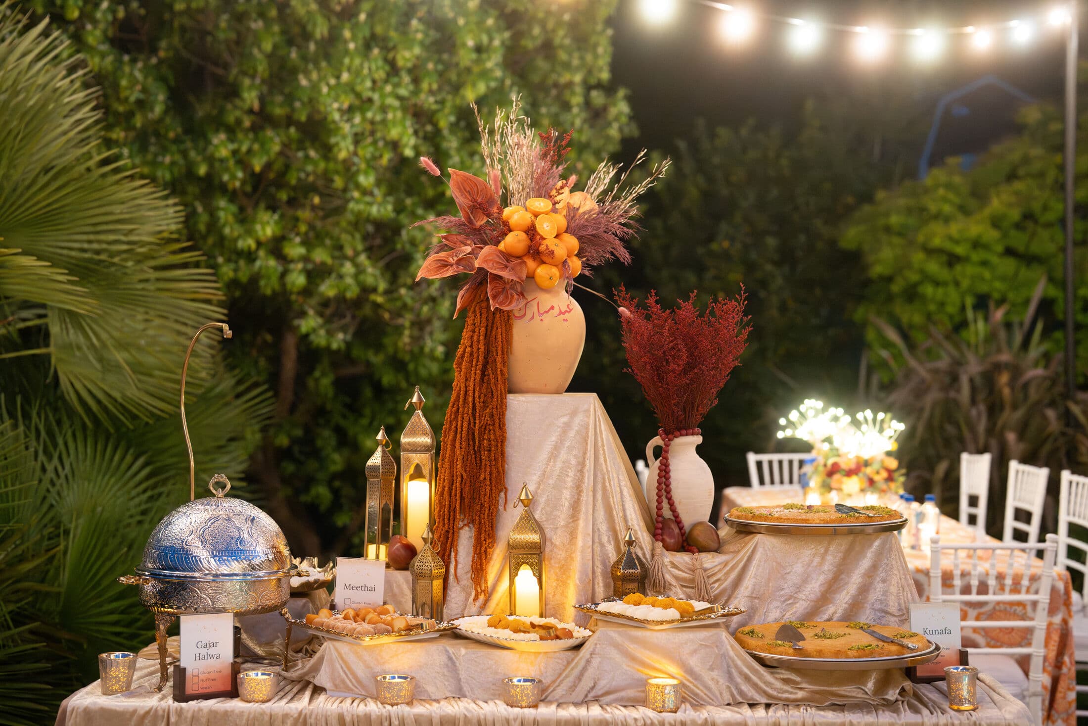 An evening garden dessert display by Memorable Moments by Ladle n Wok, styled with tall gold lanterns, a rust-and-cream floral urn, labeled desserts, and a domed cloche under string lights.