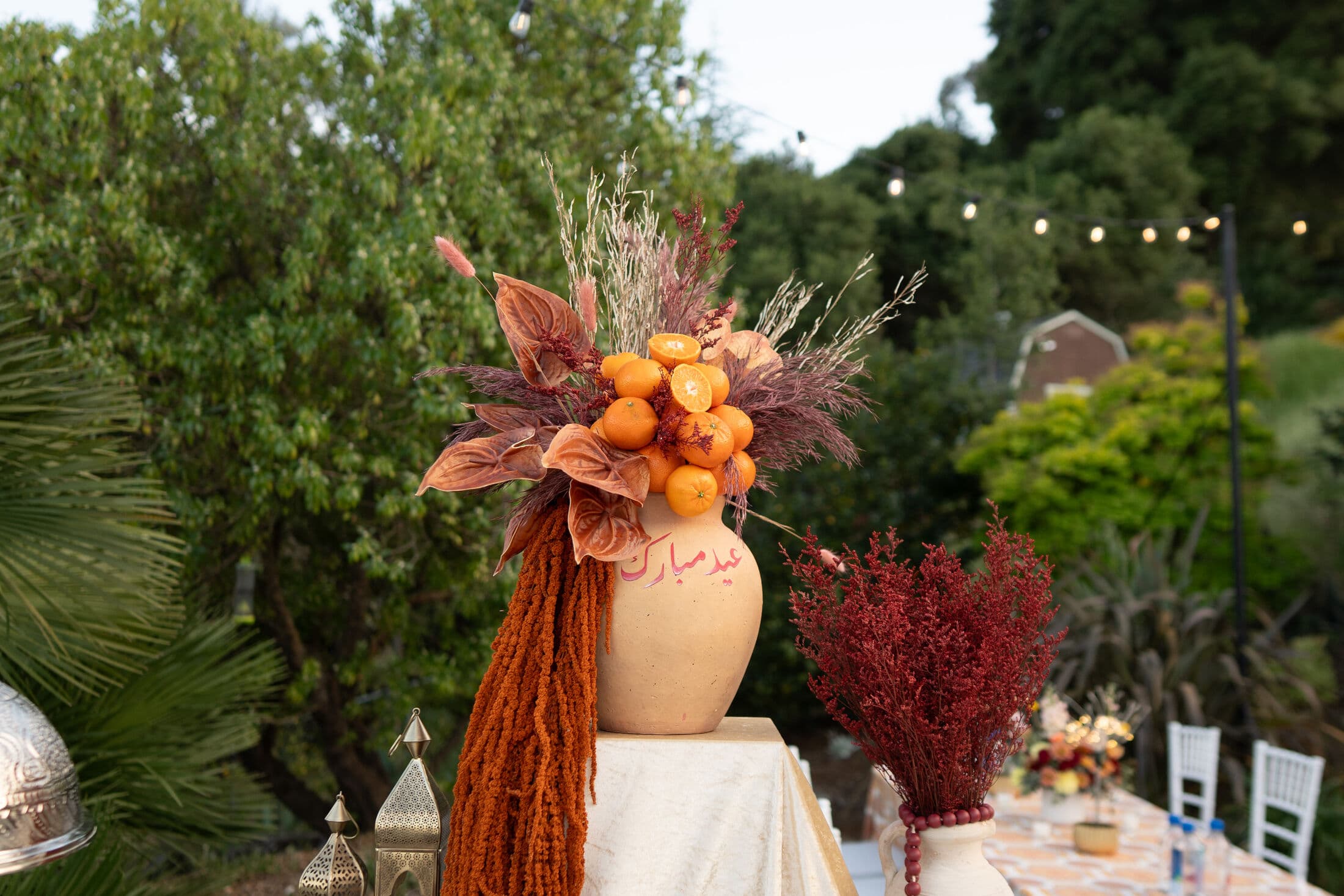A cream urn arrangement of clementines, dried rust foliage, and cascading tassels with Arabic 'Eid Mubarak' script, set on a Bay Area garden table beside a burgundy dried-flower vase.