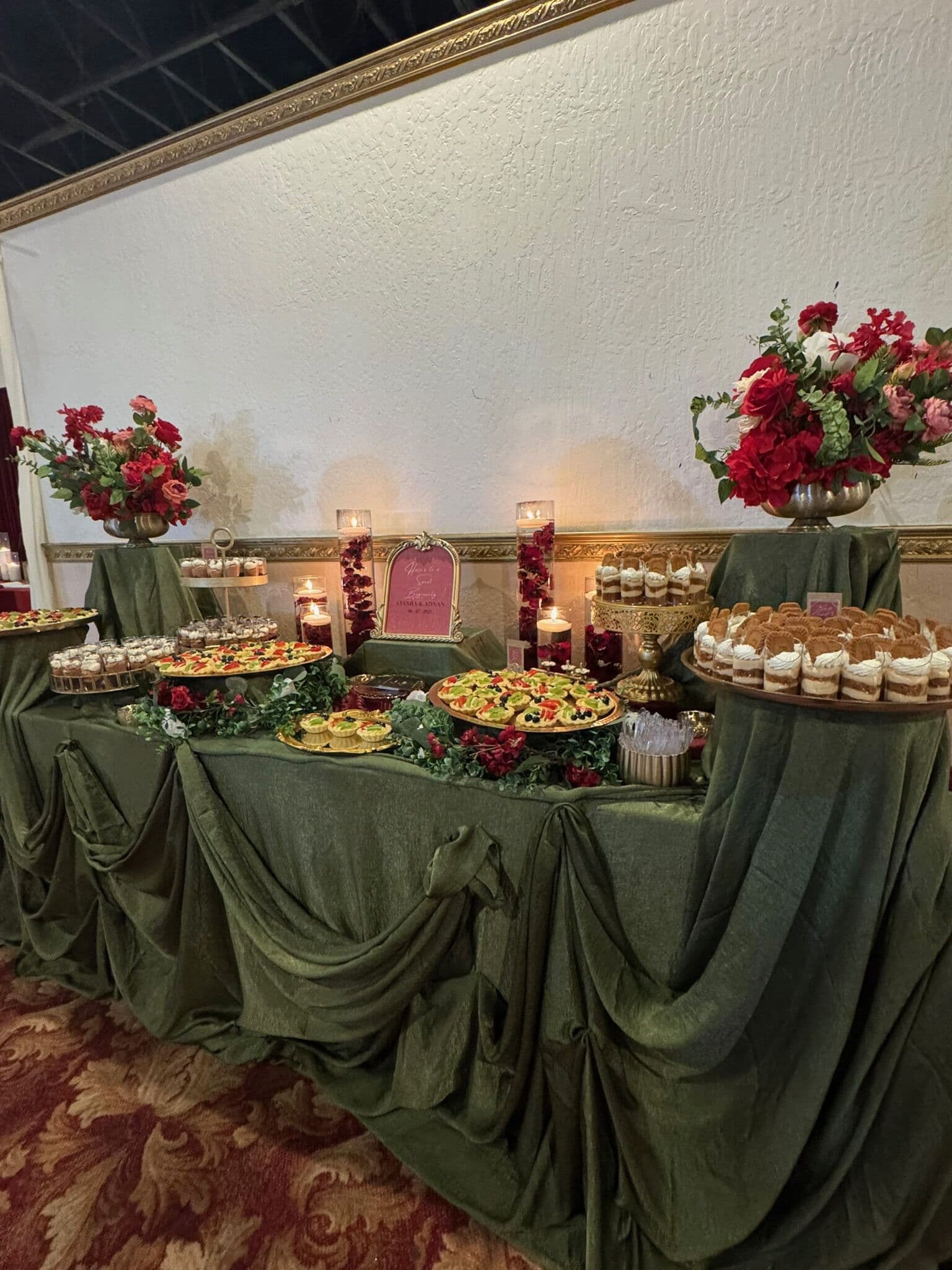 An olive-green draped dessert table with tiered cupcakes and mithai trays, flanked by tall red rose arrangements and pillar candles against a gold-framed ballroom wall.