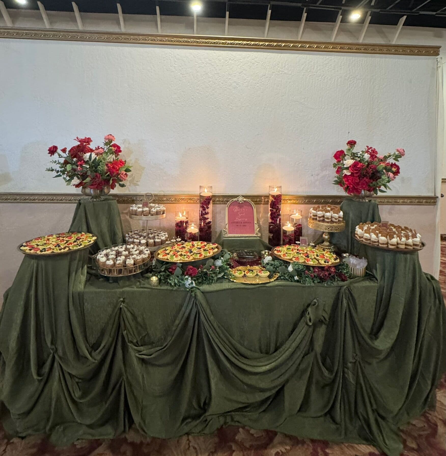 A symmetrical dessert spread on olive draping with pistachio-topped cupcakes, fruit tarts, floating-candle vases and red roses, centered on a pink arched signboard in a Bay Area ballroom.