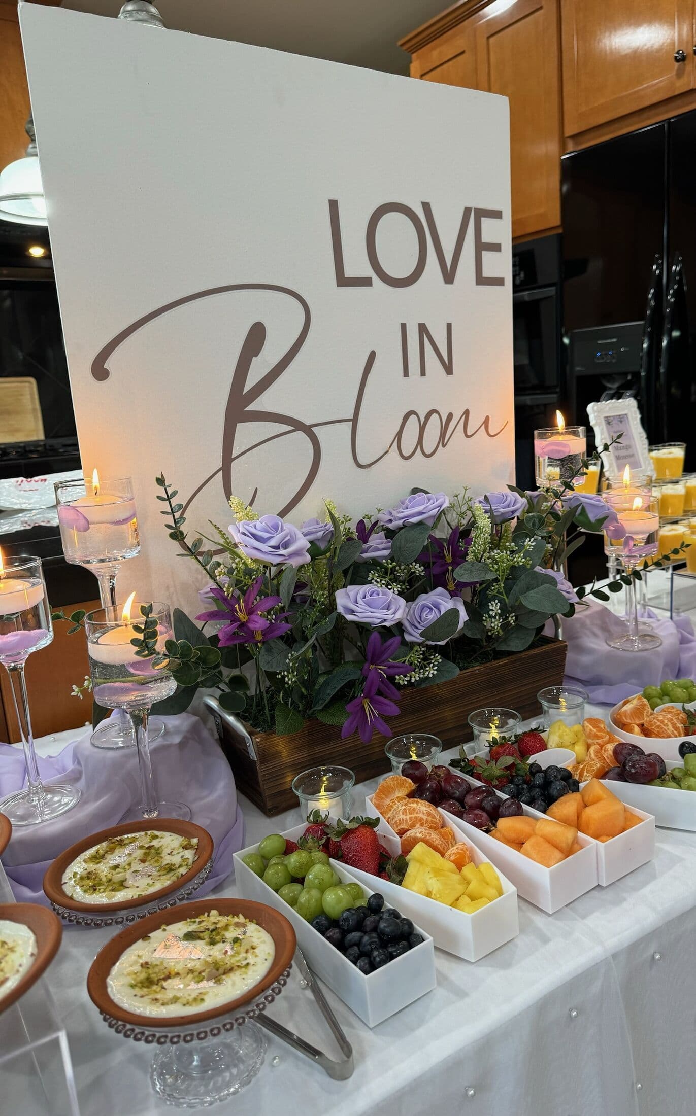 A lavender-themed dessert and fruit table with a 'Love in Bloom' sign, purple roses, mimosa glasses, and white compartment trays of melon, grapes, and berries beside small dessert cups.