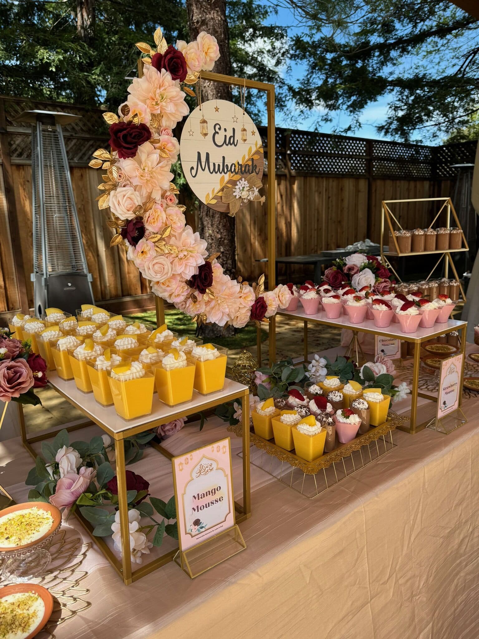Tiered gold stands of mango mousse cups topped with cream and a berry, set below a floral 'Eid Mubarak' hoop on a backyard Bay Area dessert table with a labeled mango mousse sign.