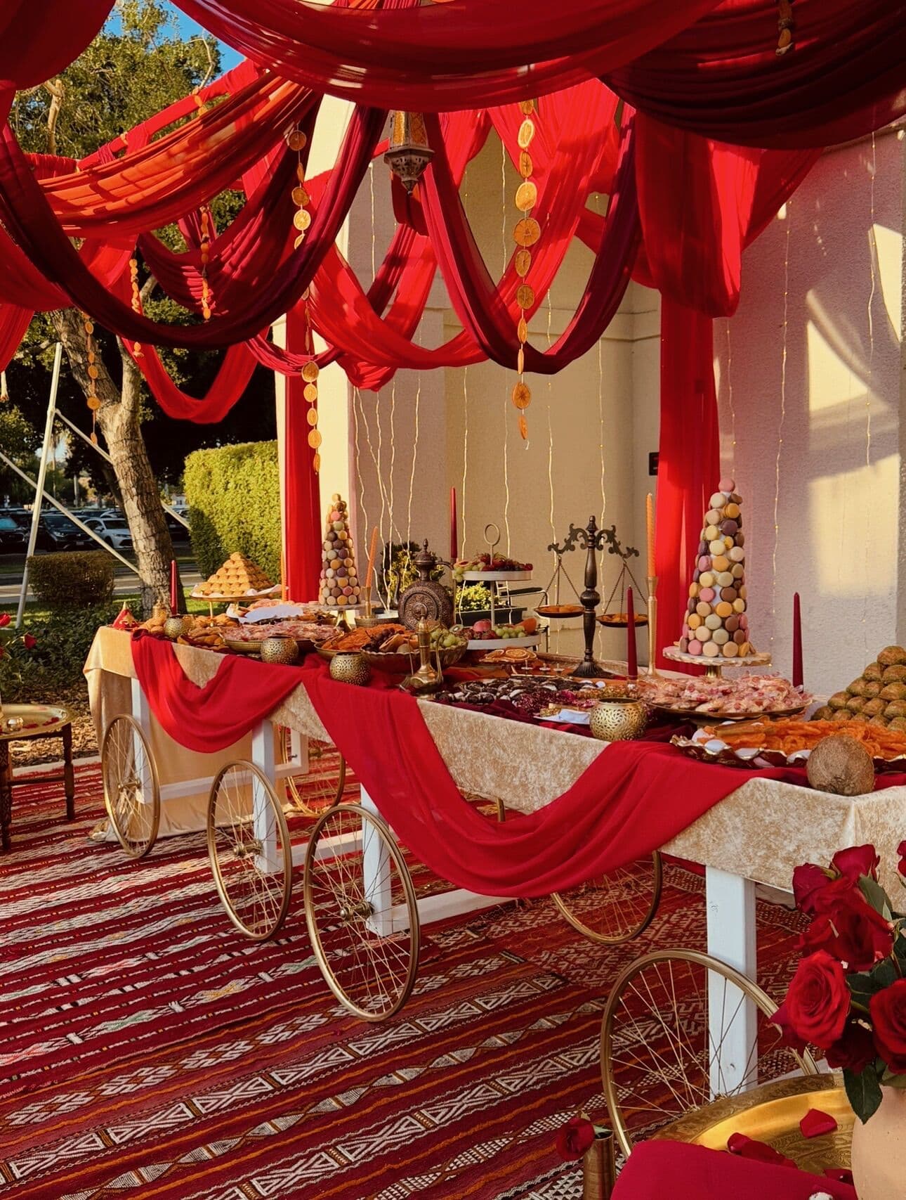 An outdoor dessert table beneath dramatic red and burgundy ceiling swags, with gold-wheeled cart tables, a macaron tower, tapered candles, and sweets platters over a woven rug.
