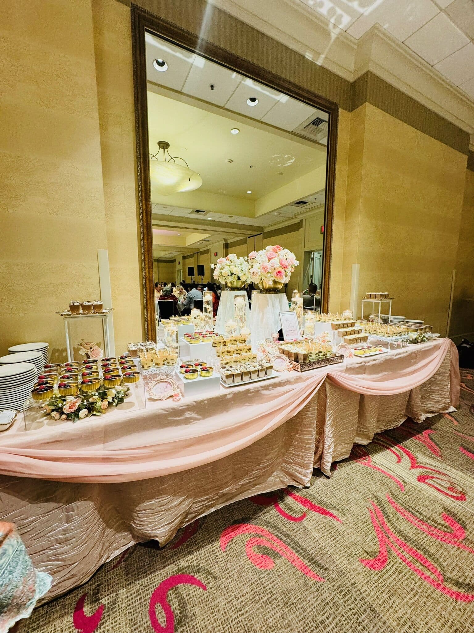 A blush-draped dessert table before a large gilt mirror, layered with dessert cups, tarts, and low pink-and-white floral centerpieces in a Bay Area hotel ballroom.