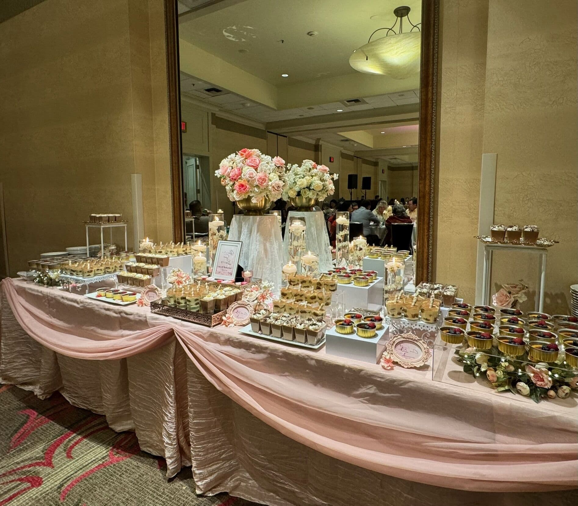 A long pale-pink draped dessert table with tiered stands of dessert cups and mini pastries, a pink-and-white rose centerpiece, and a small signboard reflected in a gold-framed mirror.