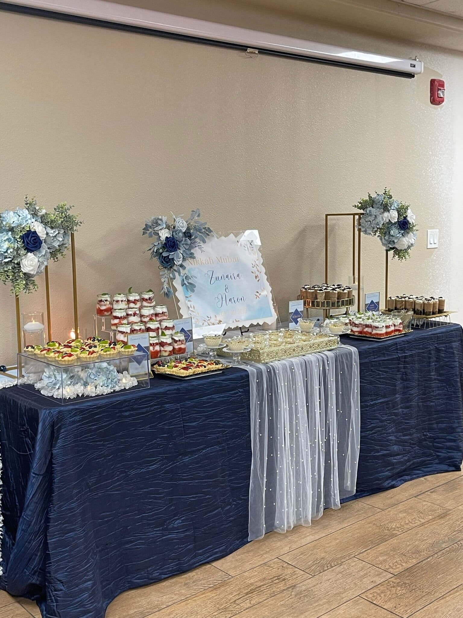 A navy-draped dessert table with a soft blue watercolor sign, dusty-blue floral arrangements on gold stands, layered dessert cups, and tarts, with a beaded sheer overlay down the center.