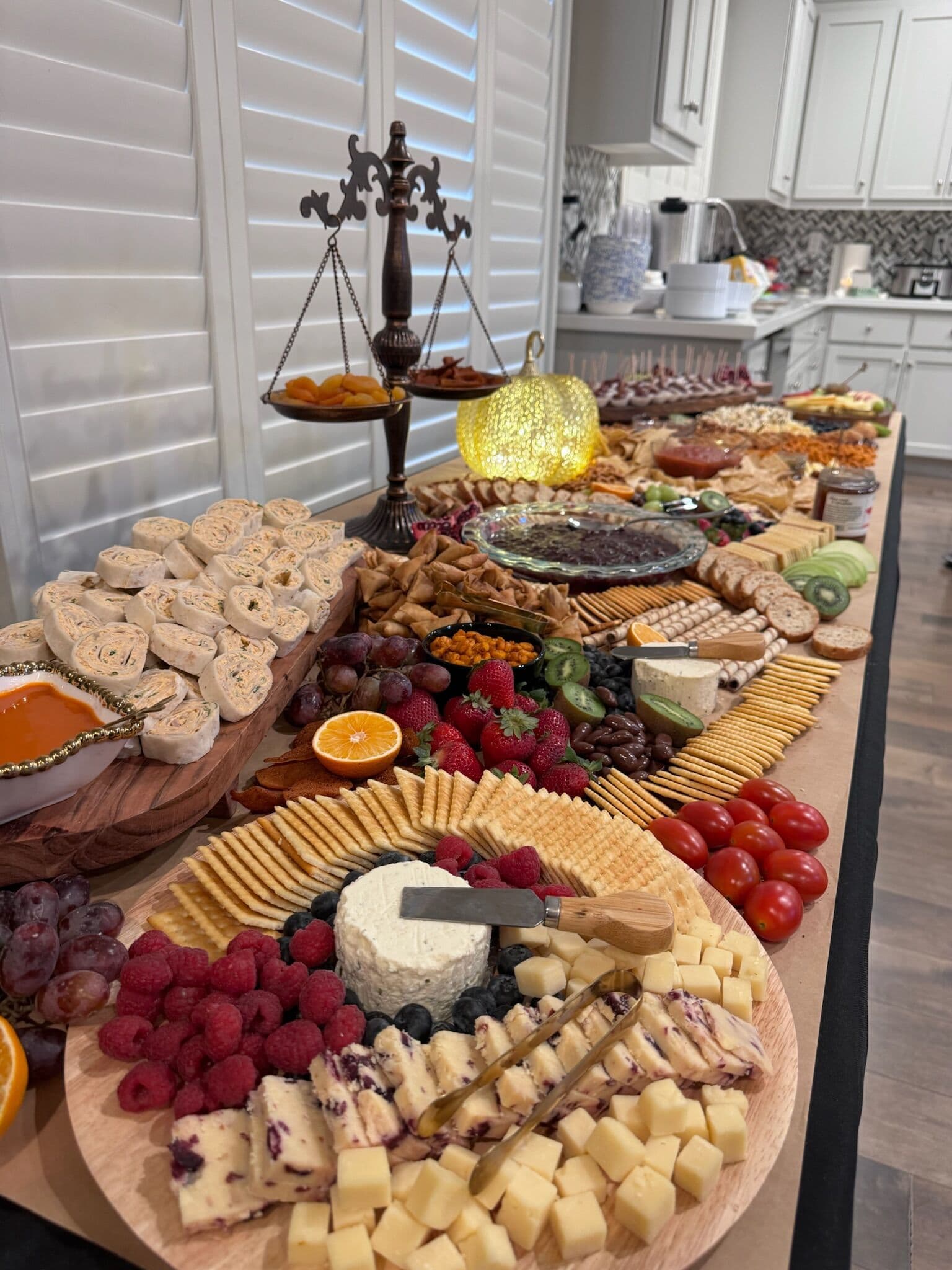 An abundant grazing and dessert board across a kitchen island with wheels of cheese, crackers, cured rolls, raspberries, grapes, and citrus, centered on a brass balance-scale prop.