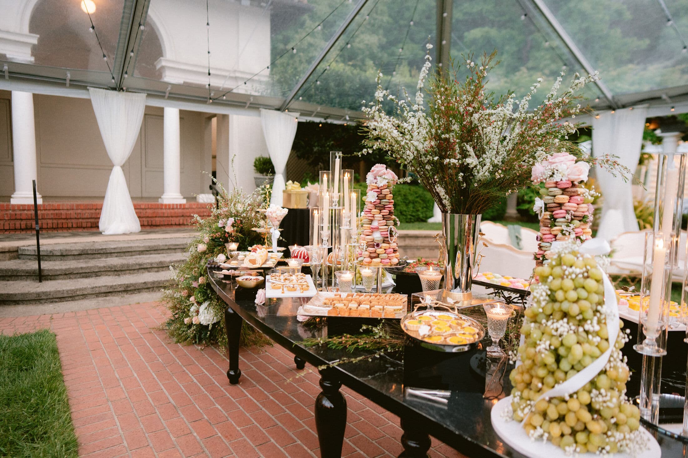 An elegant black-table dessert display under a clear garden tent with macaron towers, tiered cups, tarts, cascading grapes, and tall tapered candles amid greenery and baby's breath.